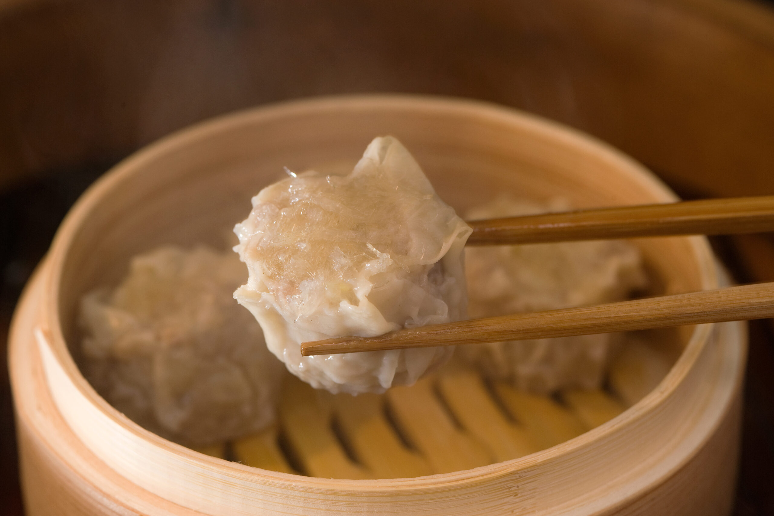 Dumpling held with chopsticks over a steaming bamboo basket.
