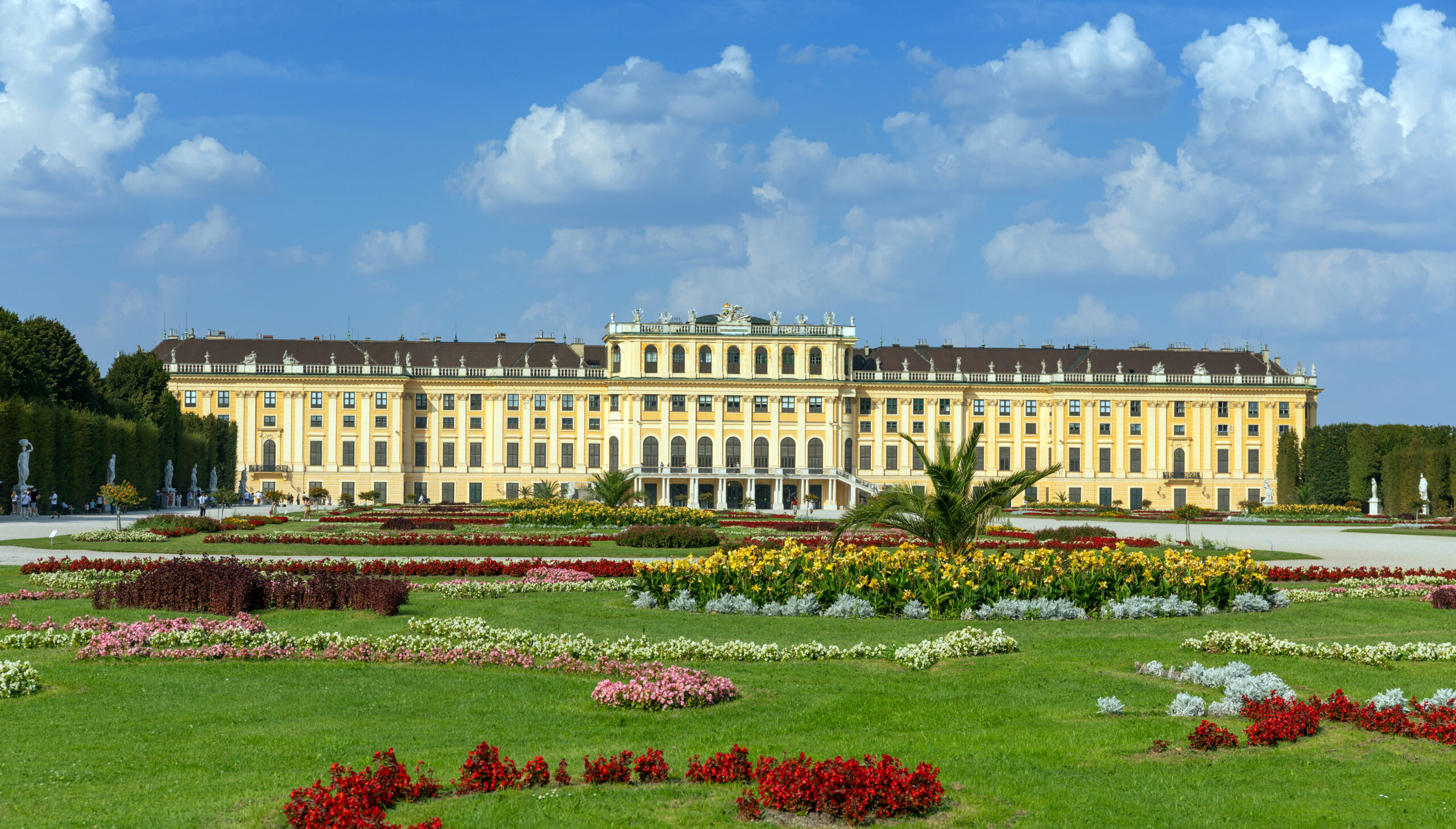 Front view of a grand yellow historic palace with symmetrical wings, ornate statues, and a formal garden of colorful flower beds under a bright blue sky.