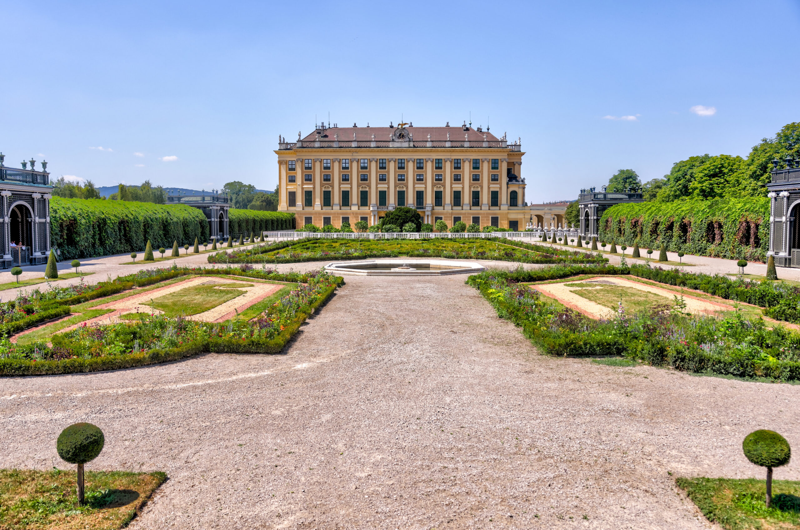 Central yellow Baroque palace with a symmetrical façade, framed by formal gardens of trimmed hedges, gravel paths, and a circular fountain in the foreground under a clear blue sky