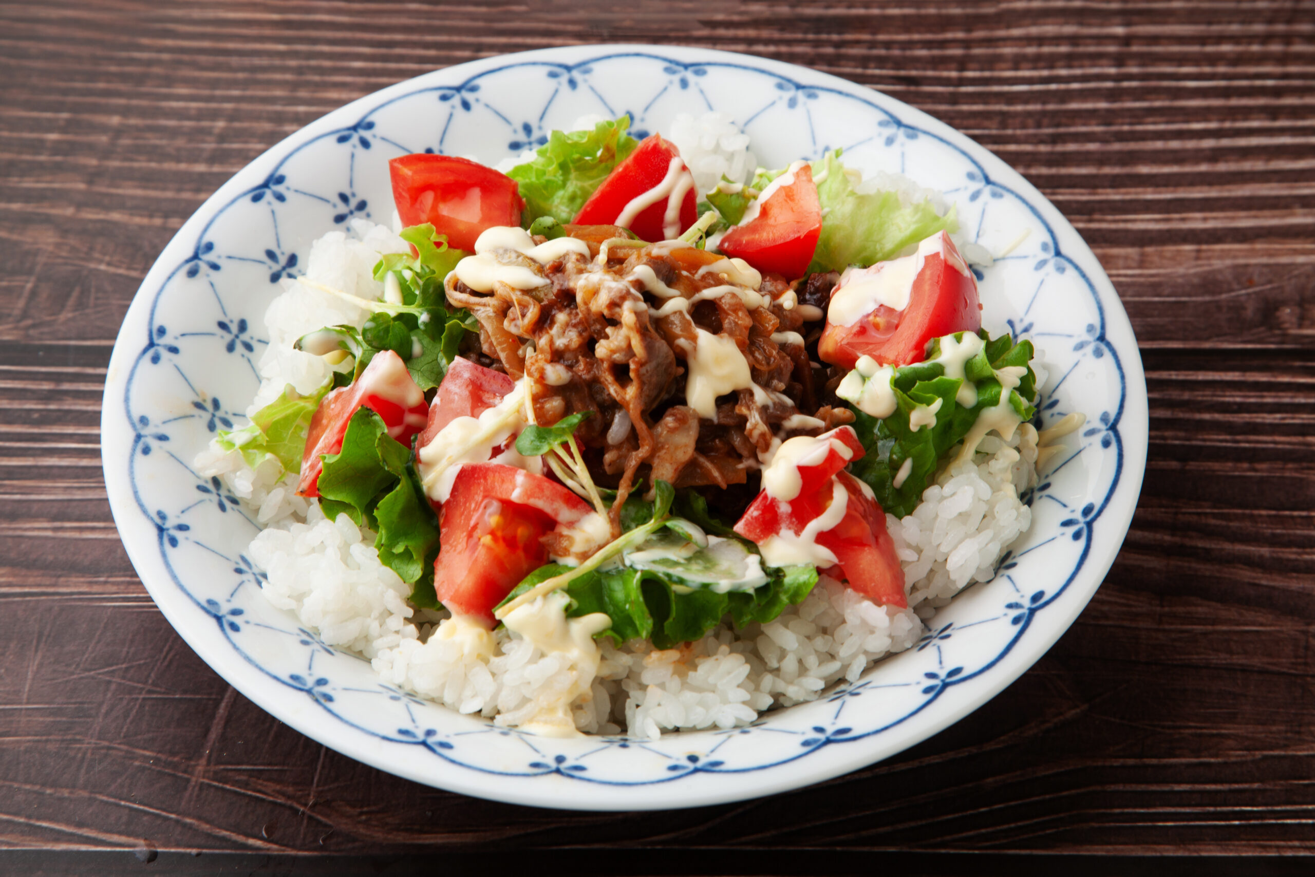 Plate of white rice with a vegetable salad and shredded beef, drizzled with creamy sauce on a blue-patterned plate atop a wooden table.