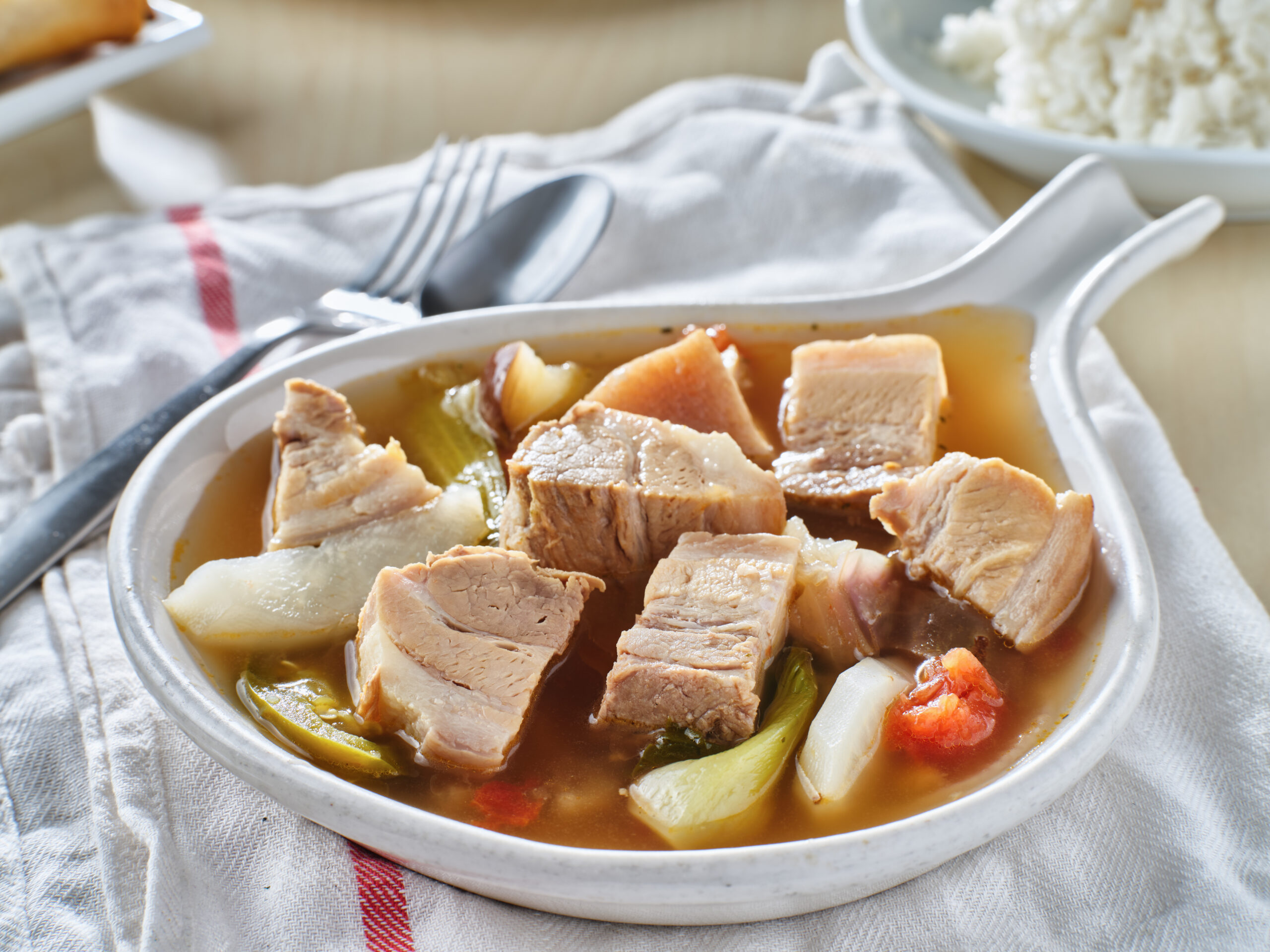 Bowl of pork and vegetable soup with chunks of pork in a light broth on a striped napkin.