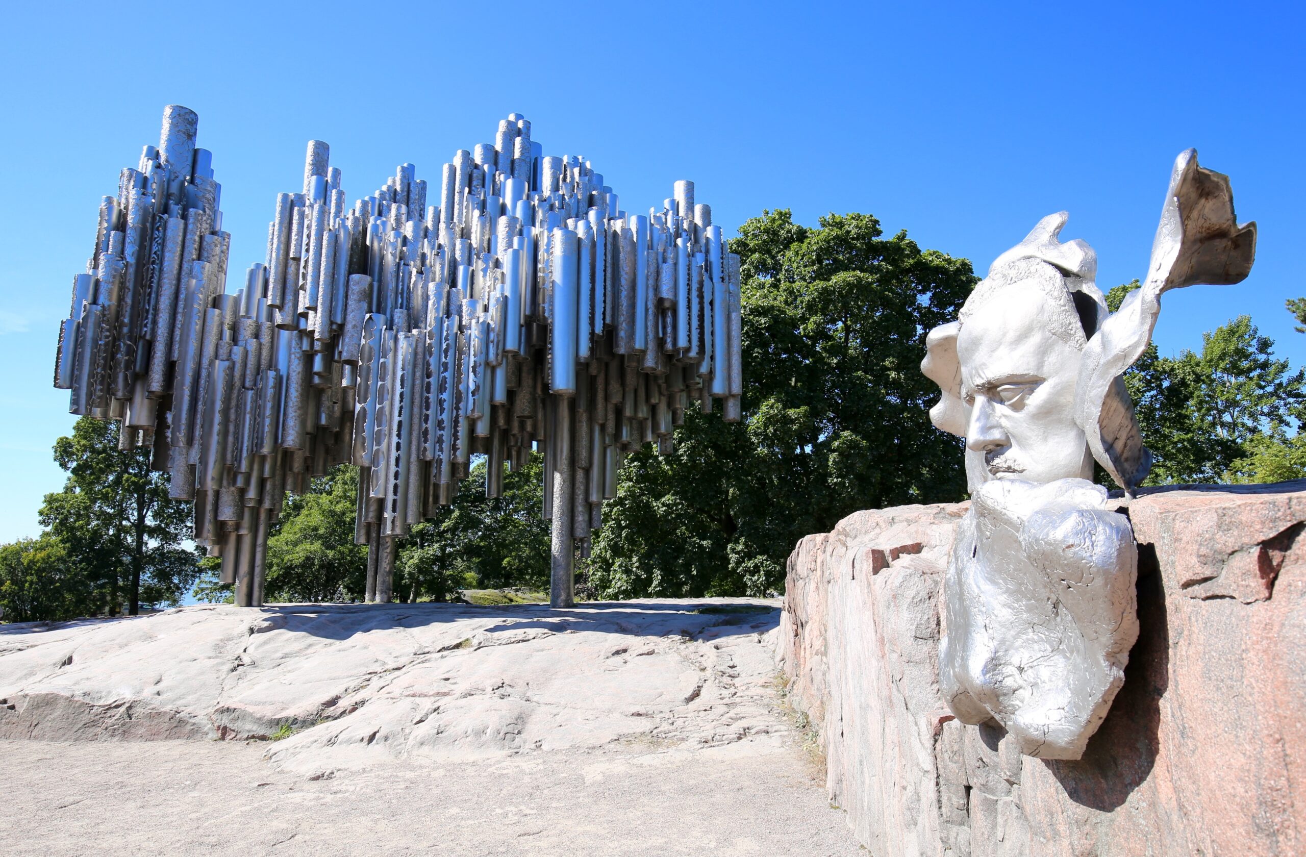 Outdoor sculpture: dense cluster of vertical metal tubes on the left and a silver bust with a winged/helmeted form on the right, blue sky behind.
