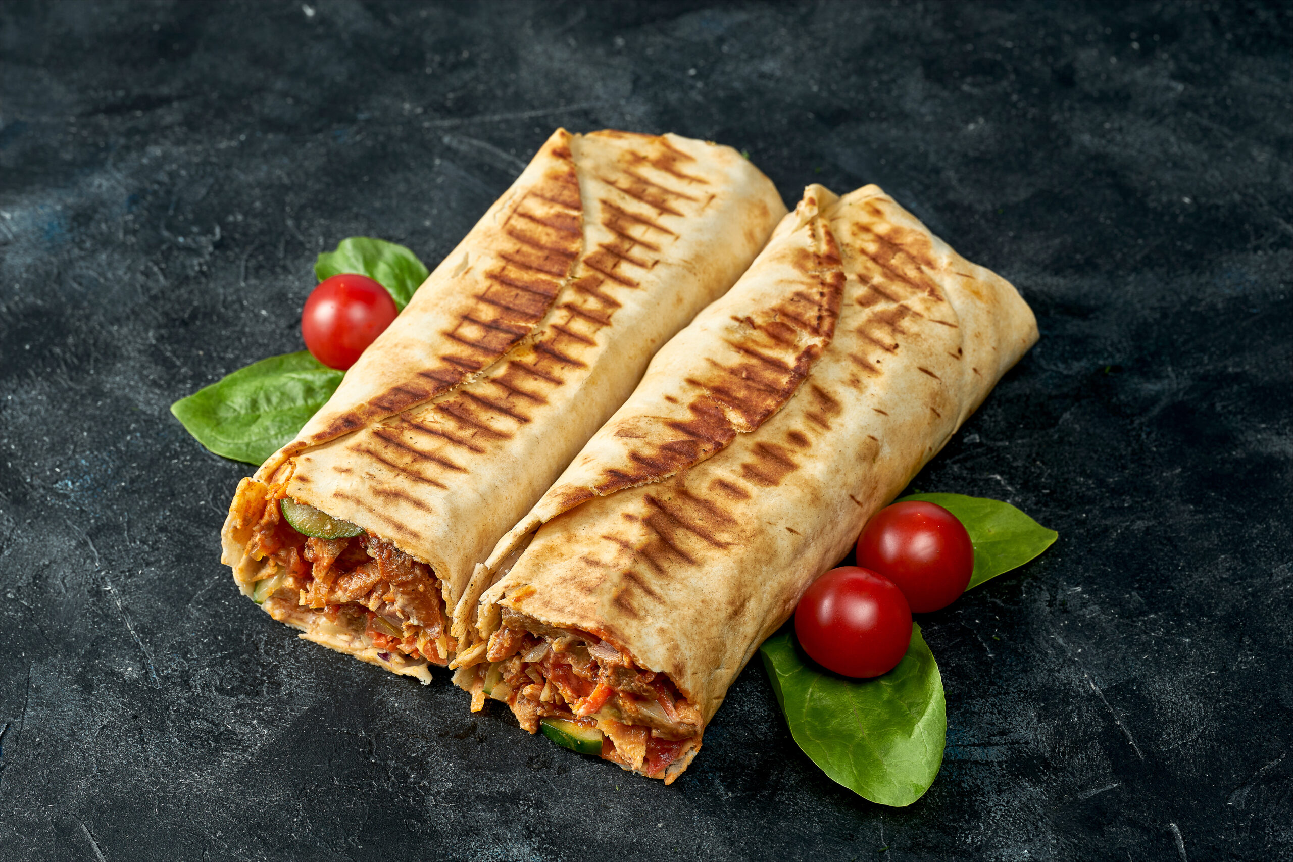 Two grilled burritos sliced open to reveal seasoned meat and vegetables, with cherry tomatoes and greens on a dark surface
