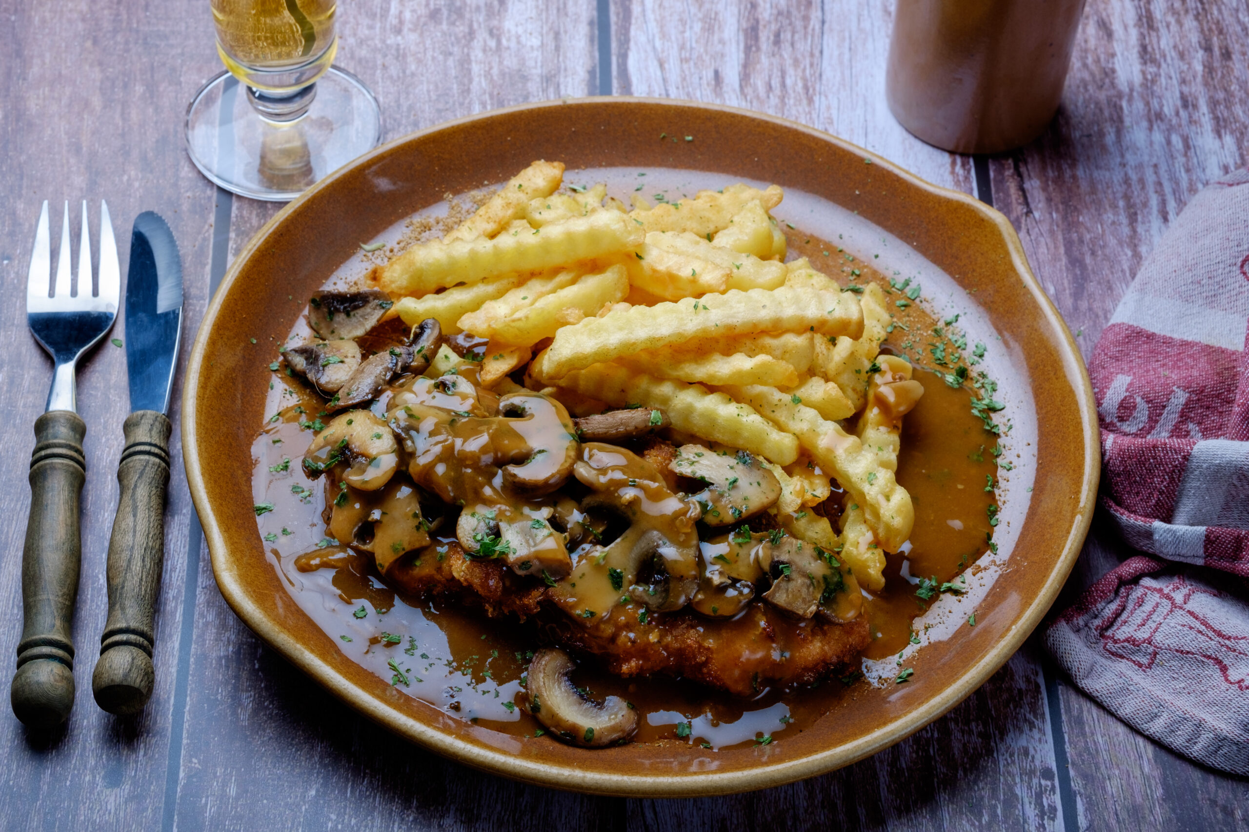 Plate of crinkle-cut fries with mushroom gravy and sautéed mushrooms, garnished with parsley on a brown ceramic plate; rustic table setting with cutlery nearby and a wine glass nearby.