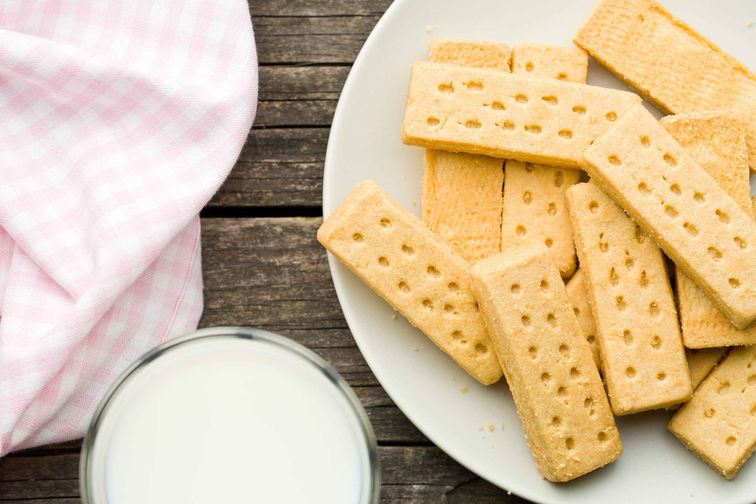 Plate with several rectangular shortbread biscuits on a white plate, next to a glass of milk and a pink checkered cloth on a wooden table.