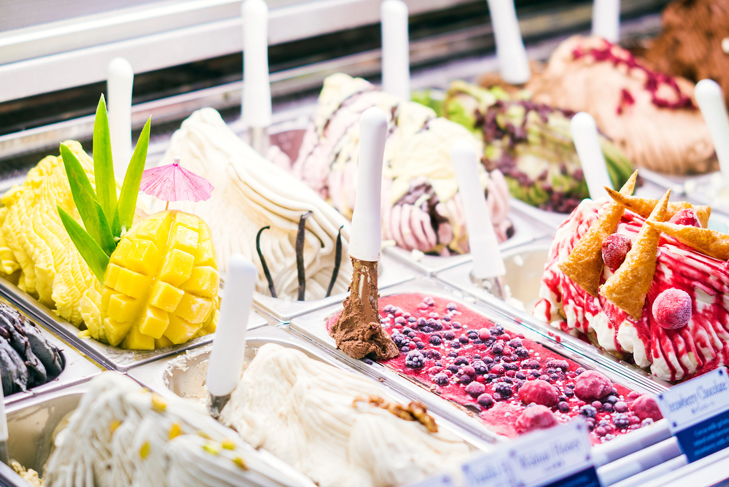 Assorted ice cream scoops with fruit toppings in a glass case, including mango chunks and berries