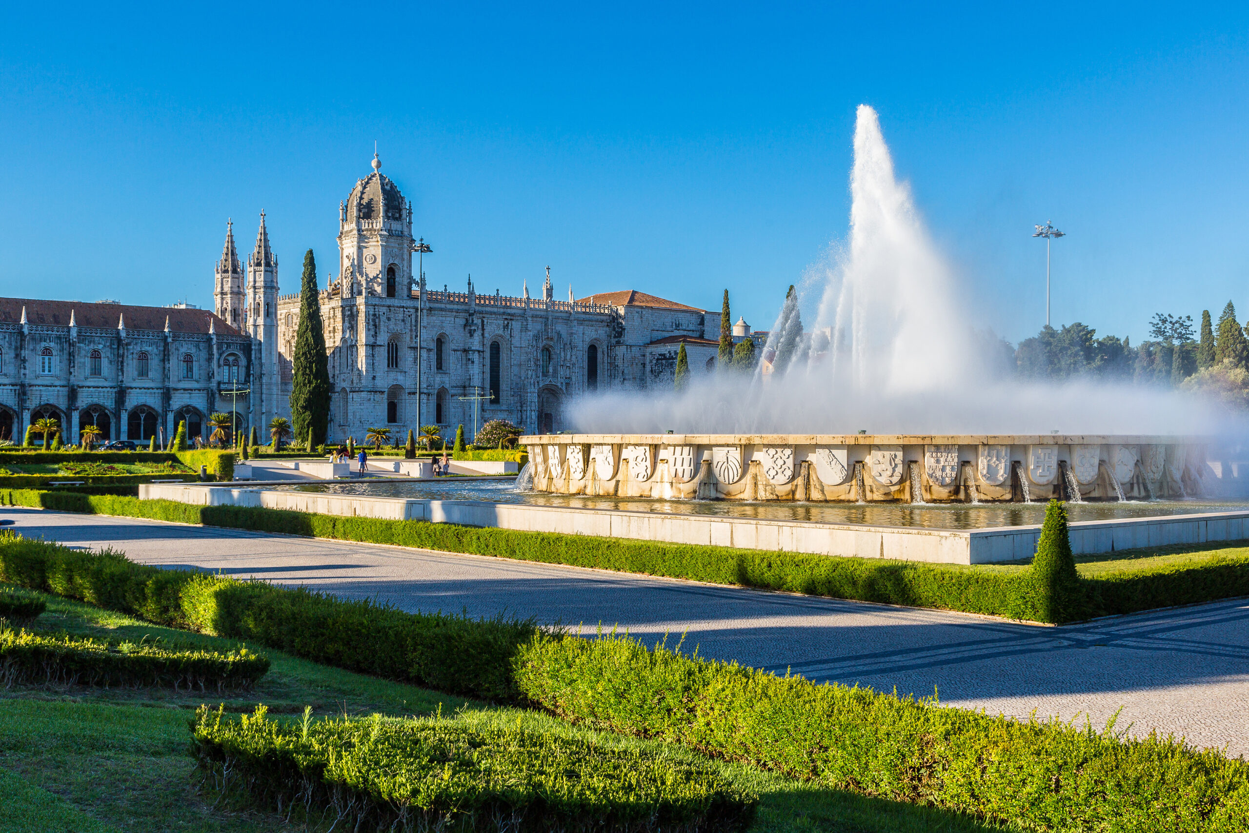 Stone historic building with multiple towers and ornate facade, facing a large fountain and formal gardens under a bright blue sky.