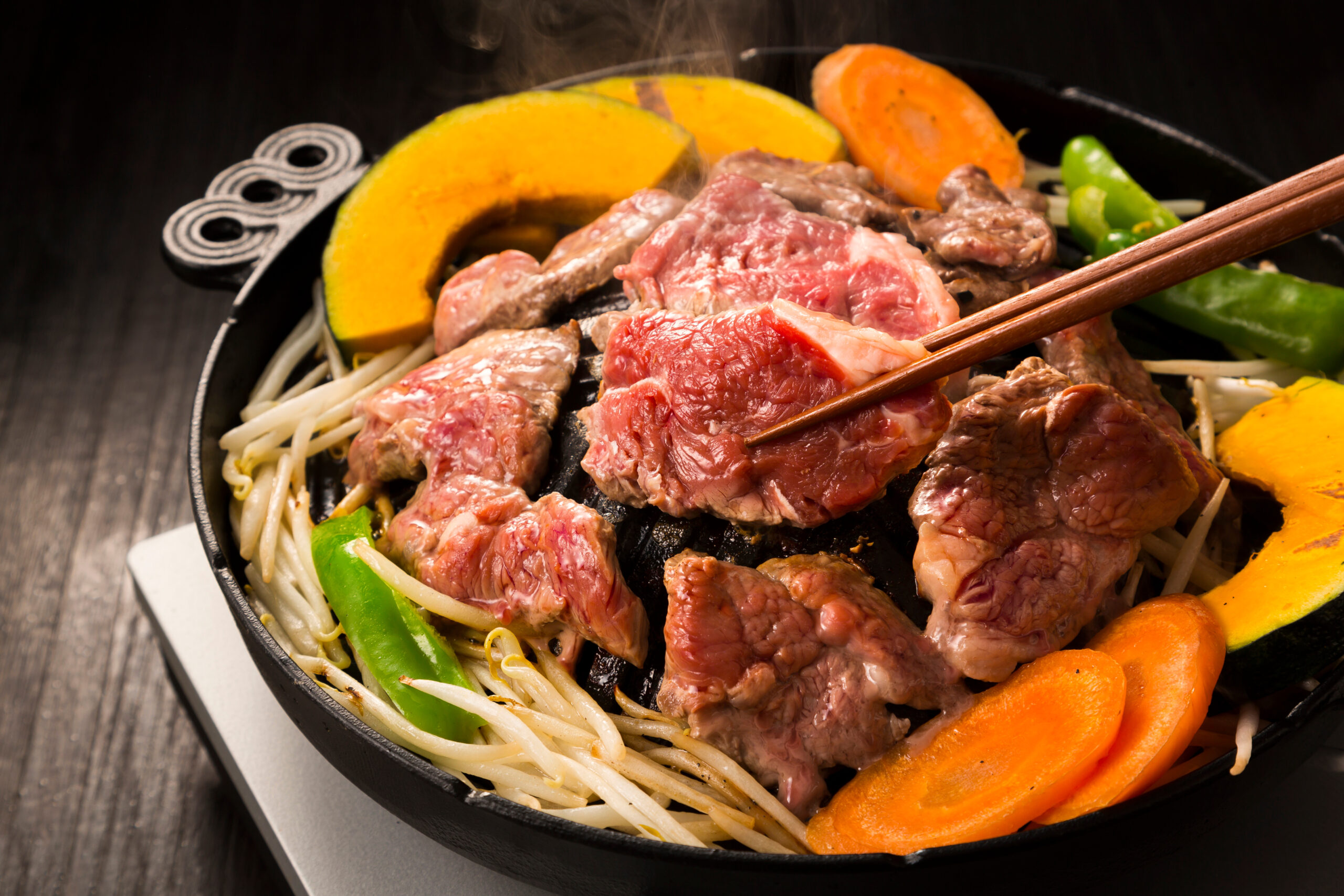 Sliced beef and vegetables cooking in a skillet with noodles and orange squash, chopsticks resting on the pan edge.