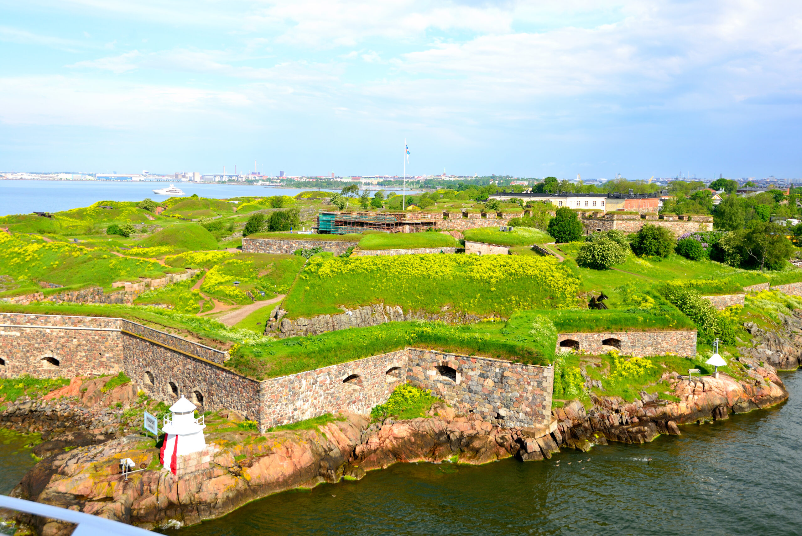 Stone fortress walls along a rocky shoreline with a white-and-red lighthouse in the foreground and green hills beyond.