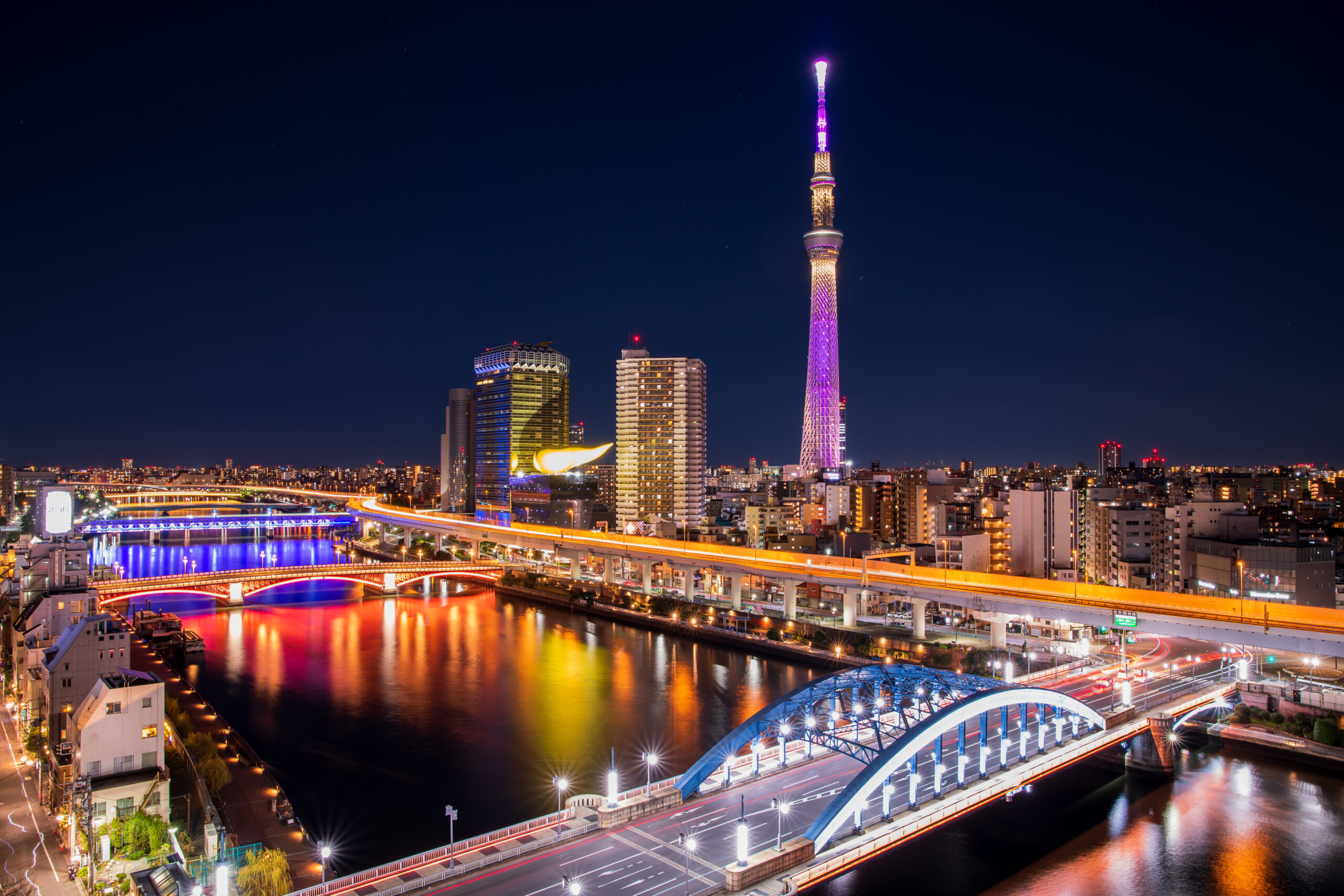 Tokyo skyline at night with a tall, purple-lit tower, illuminated buildings, and bridges reflecting in the river