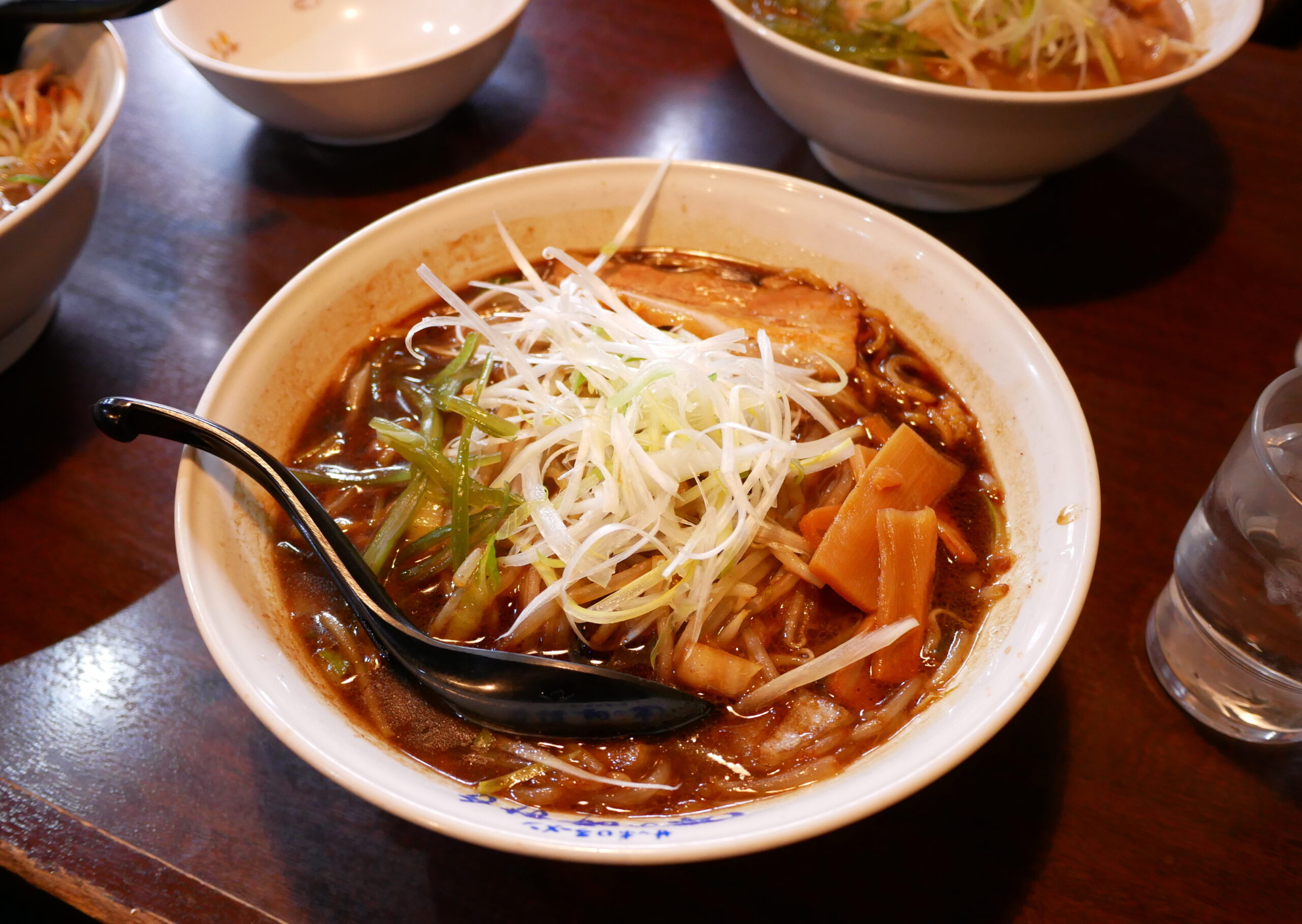 Bowl of ramen with pork slices, shredded white onions, and green onions in a spicy-looking broth on a dark wooden table.