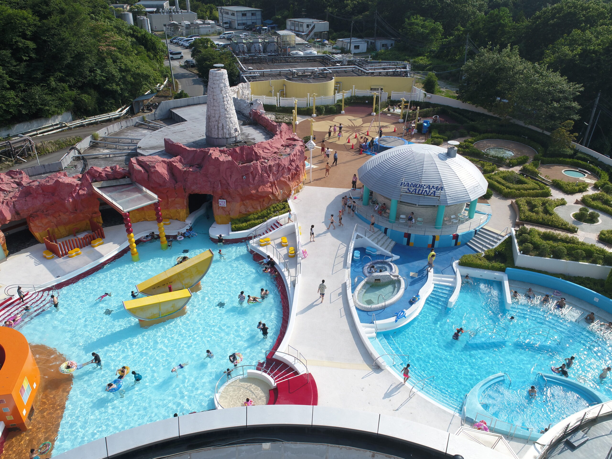 Aerial view of a colorful outdoor water park with slides, pools, and a rocky red cliff structure with people enjoying the water.