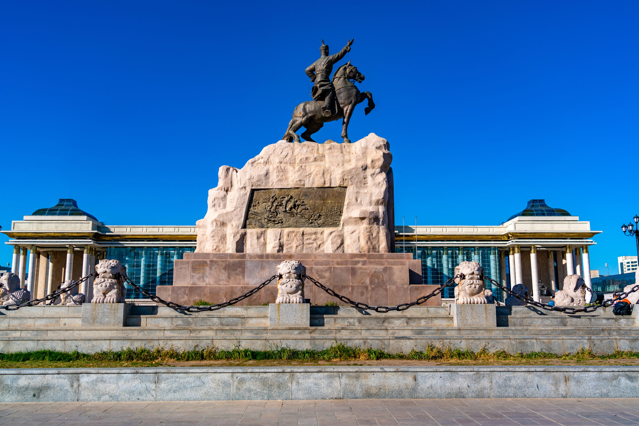 Bronze equestrian statue of a rider on a horse atop a tall stone pedestal with a bronze relief plaque below, set in a plaza under a blue sky.