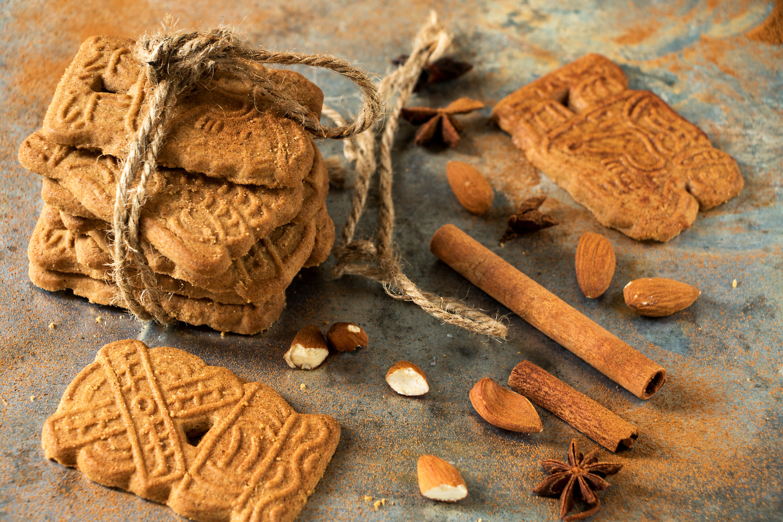 Stack of round, embossed cookies tied with rough twine on a rustic blue surface, with almonds, cinnamon sticks and star anise nearby.