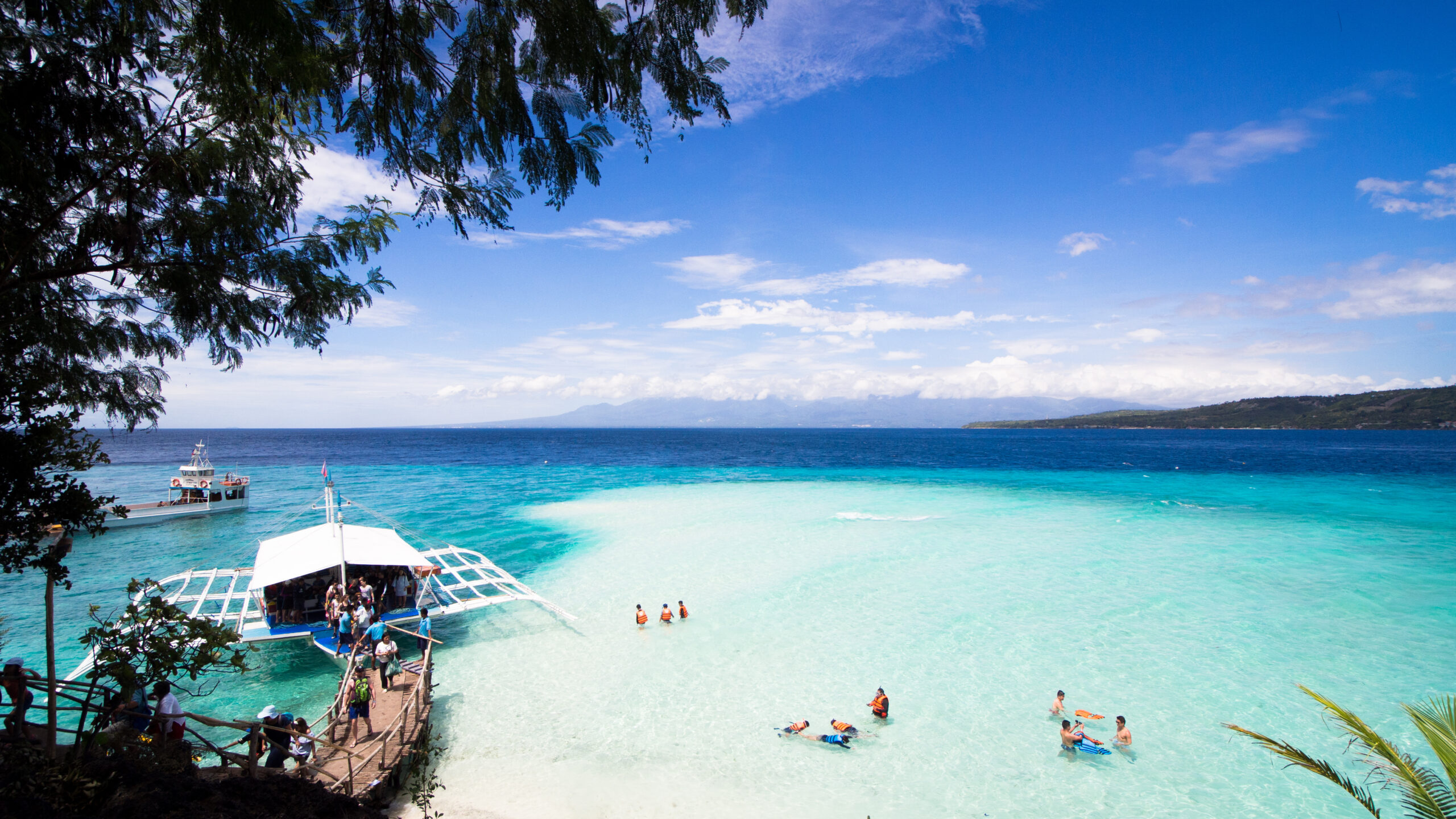 Turquoise shallow water by a beach with a wooden ramp, people wearing life jackets, boats anchored nearby, and tree branches framing the scene.