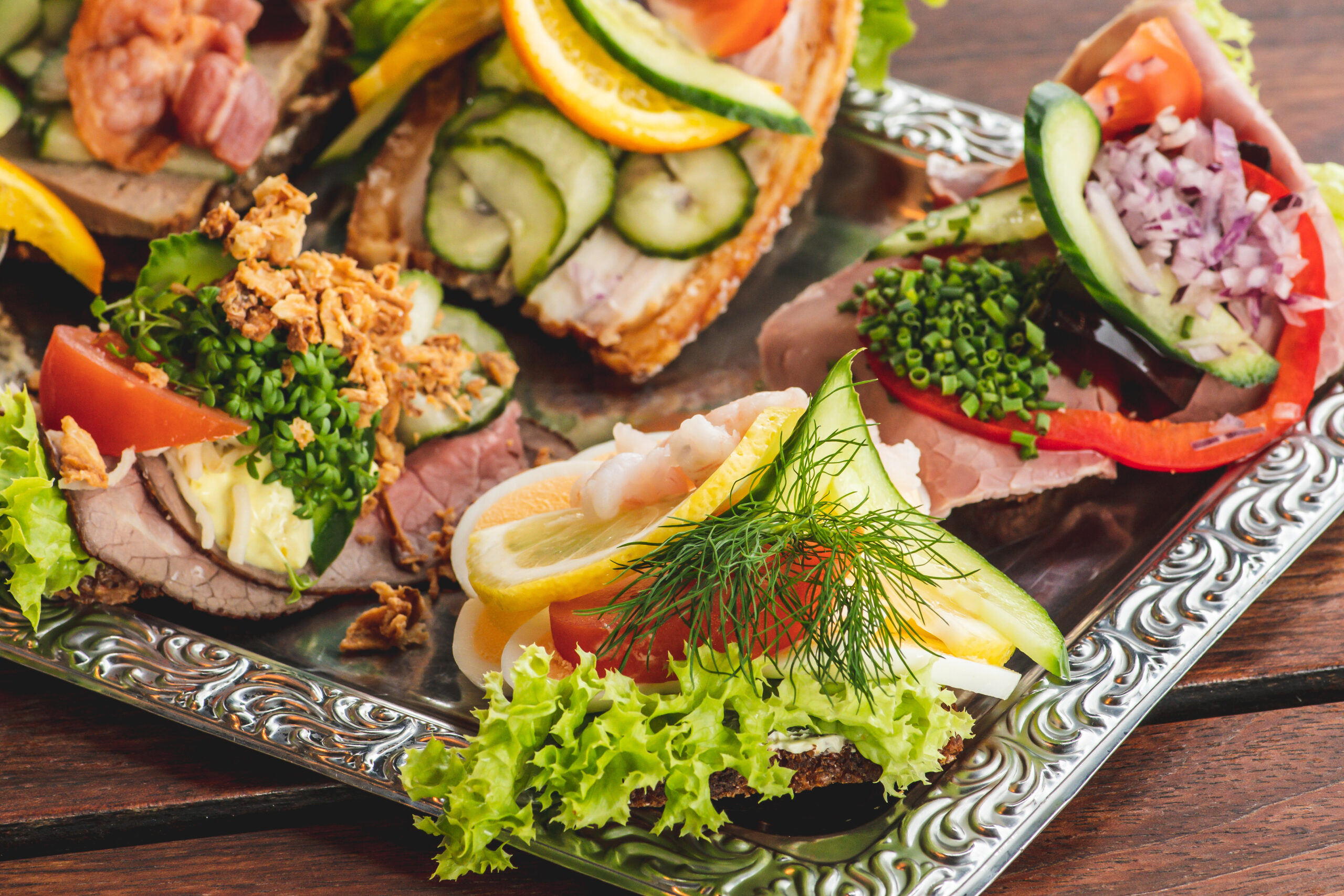 Assorted open-faced sandwiches with cucumber, tomato, dill, and greens on a decorative silver platter.