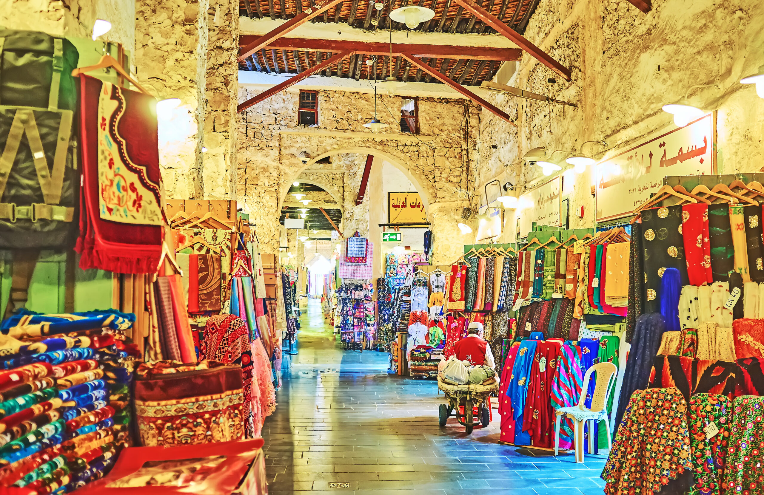 Colorful fabric stalls line a stone-walled marketplace arcade, with vibrant scarves and clothing on display under warm lighting.
