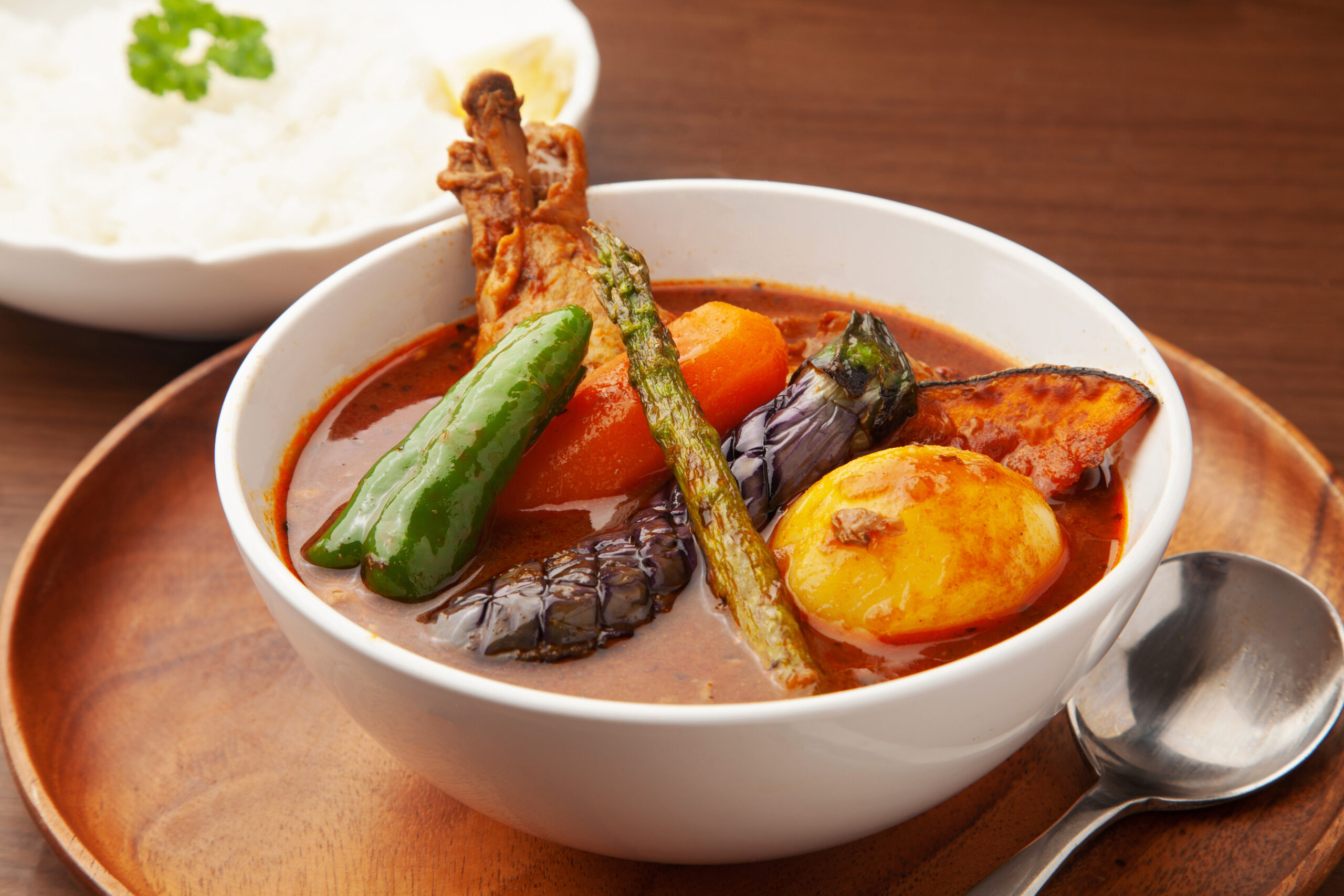 Bowl of red curry with vegetables (green pepper, carrot, eggplant, potato) and a bone-in meat piece on a wooden tray with rice in the background.