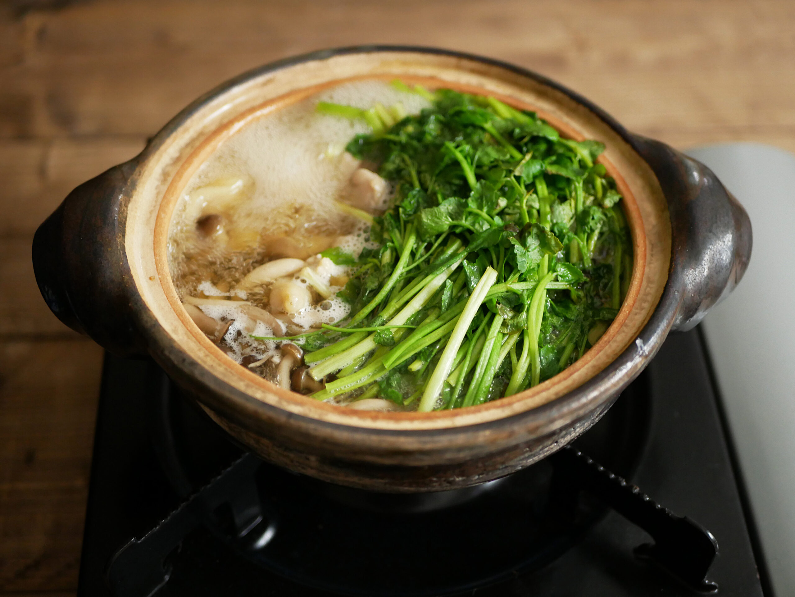 Clay pot on a stove with bubbling broth and a pile of leafy greens and mushrooms inside.