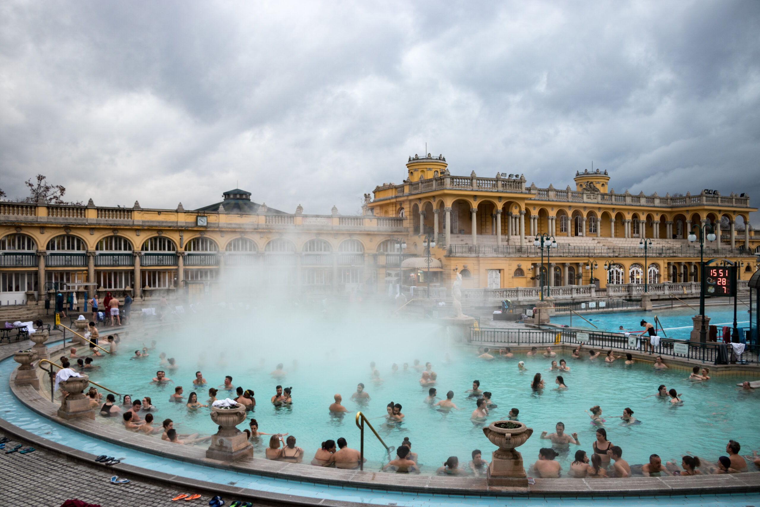 Visitors soak in a large outdoor thermal pool as steam rises, with a yellow arched historic building in the background.