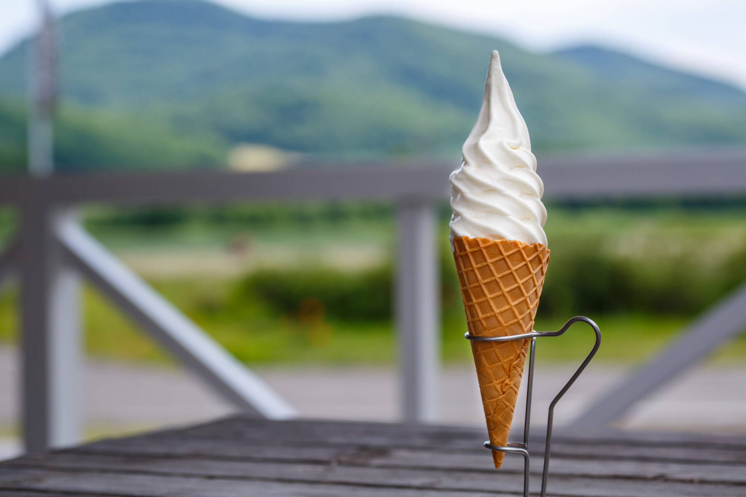 Close-up of a vanilla soft-serve ice cream cone in a waffle cone, held in a stand with a blurred outdoor mountain backdrop.