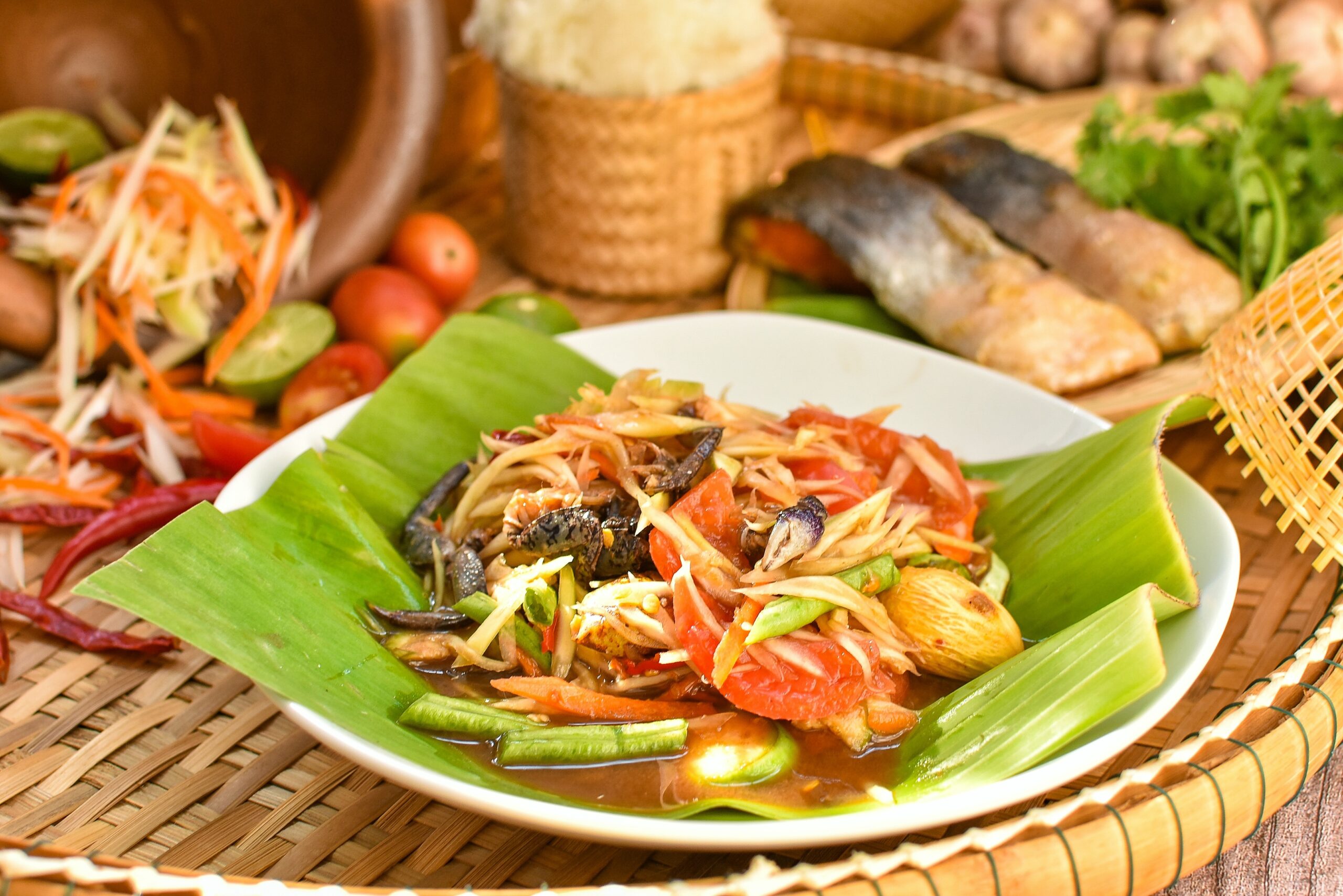 Plate of colorful papaya salad with tomatoes and carrots on a banana leaf in a white bowl, with a woven table mat.