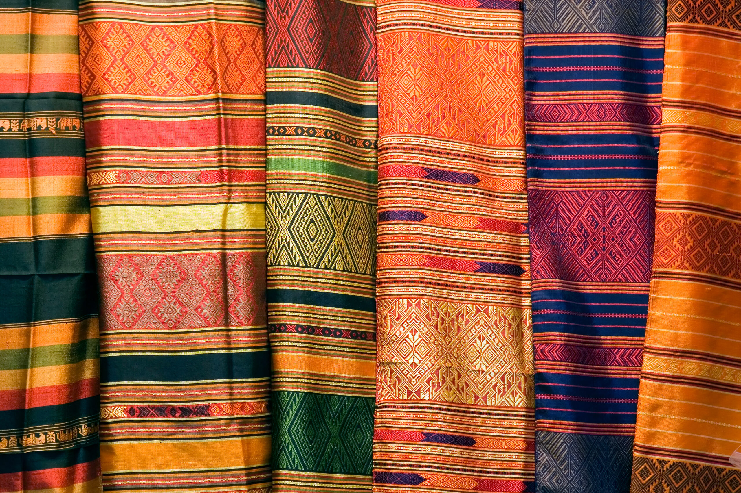 Colorful woven textiles with stripes and geometric patterns in orange, red, green, and blue, displayed in vertical rolls.