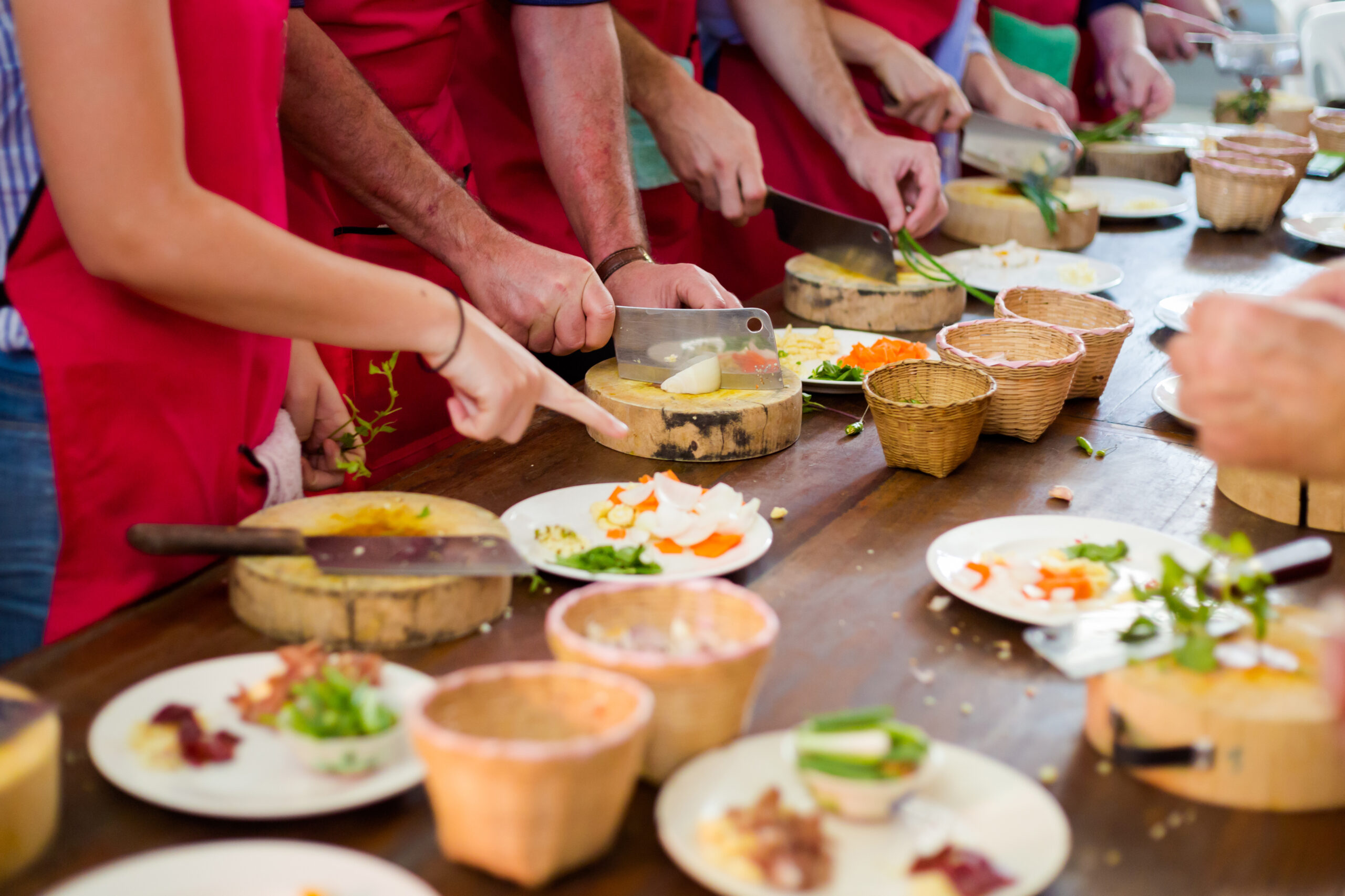 People in red aprons slice vegetables along a long wooden table, preparing ingredients.