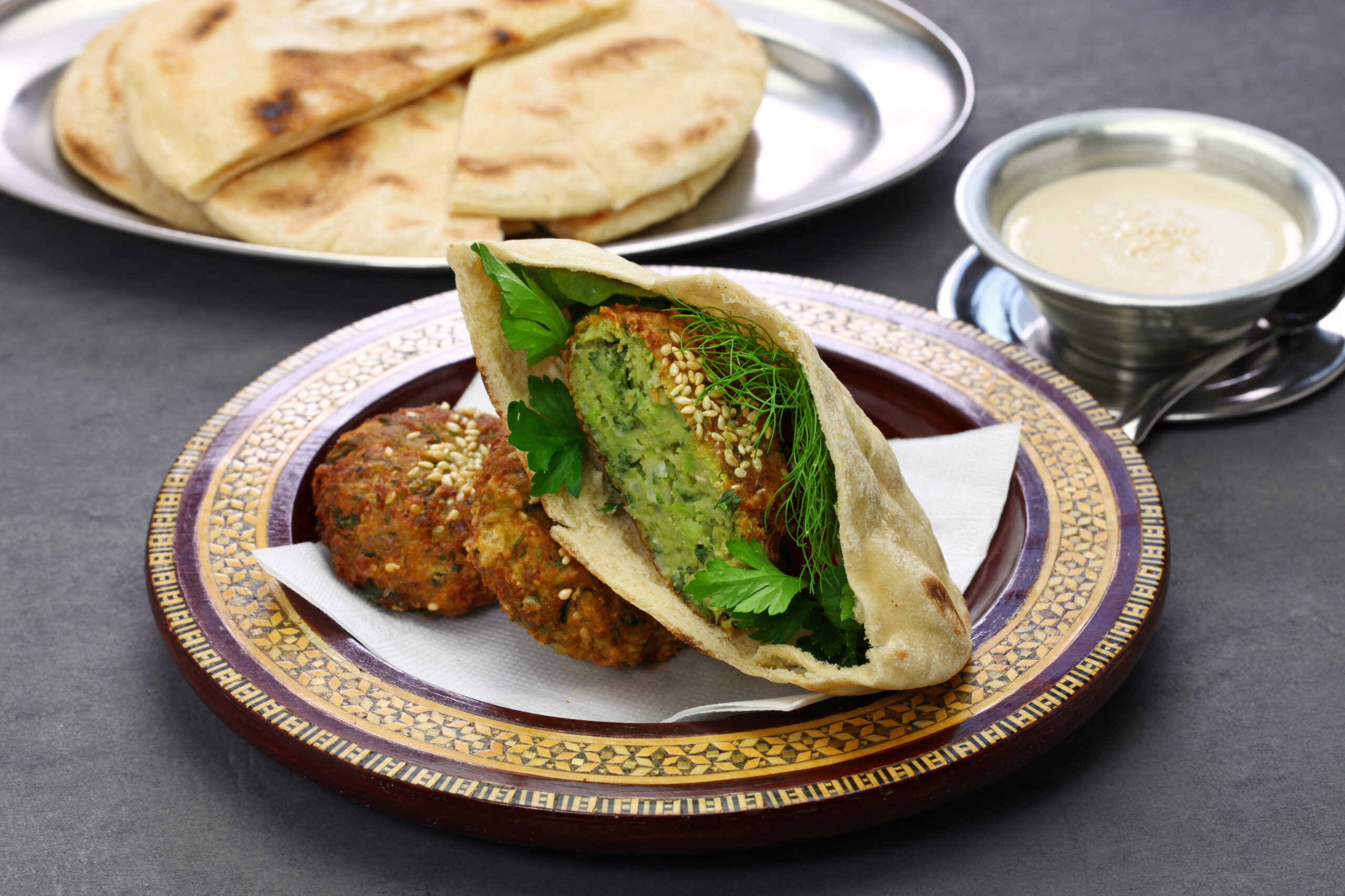 Falafel wrap opened to show green herb filling and sesame seeds on a decorative plate with naan and a creamy sauce in the background