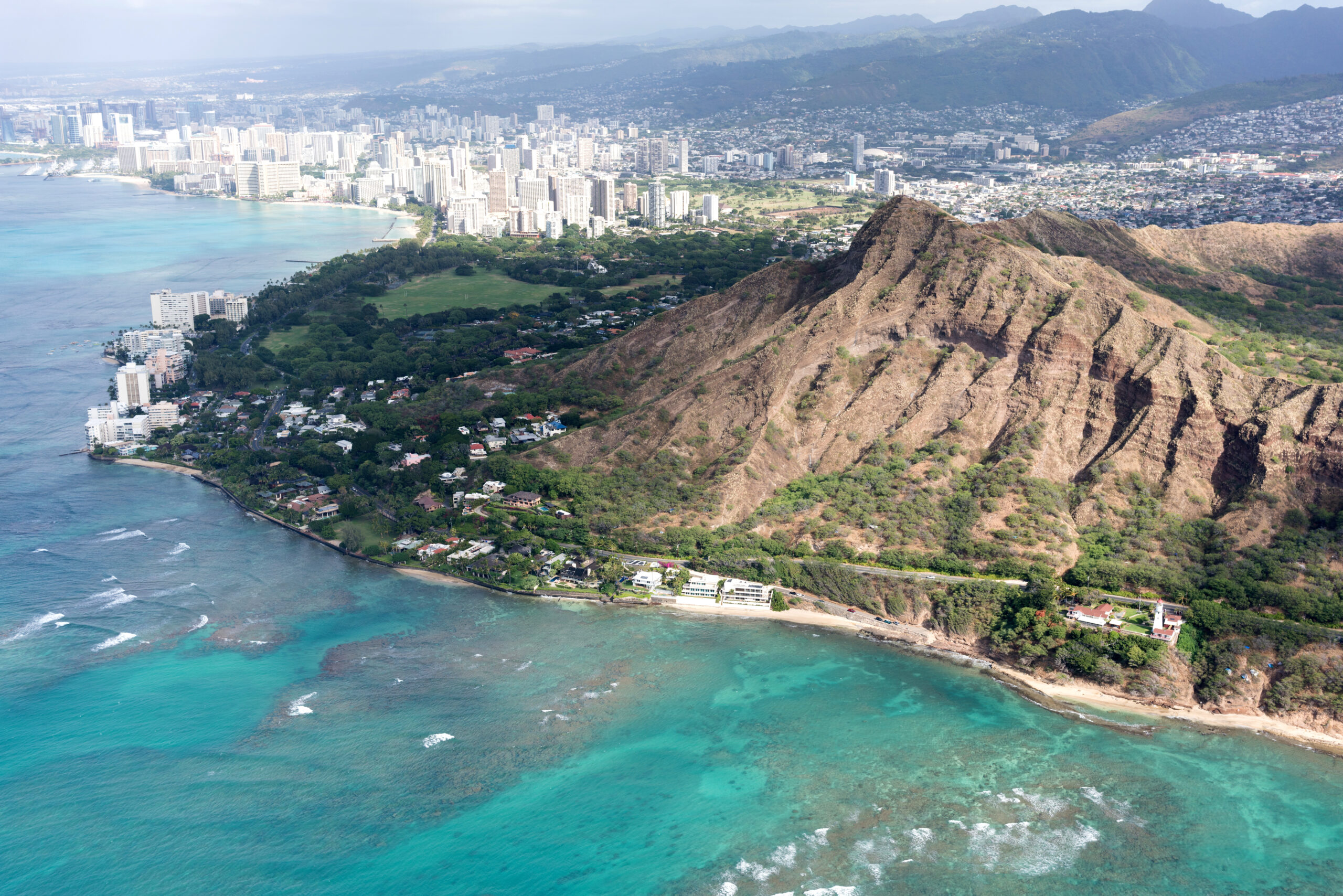 Aerial view of a tropical coastline with turquoise water, a dense city along the shore, and rugged hills in the foreground.