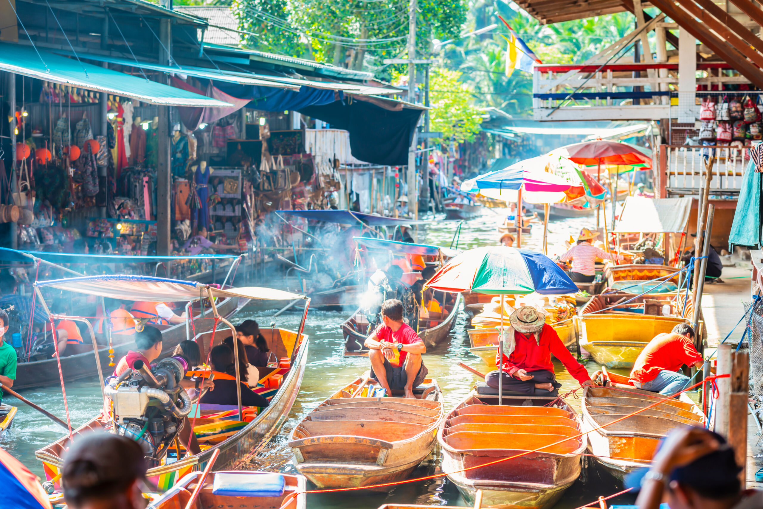 Busy floating market along a canal with colorful boats and stalls on both sides, vendors and spectators under umbrellas and awnings.