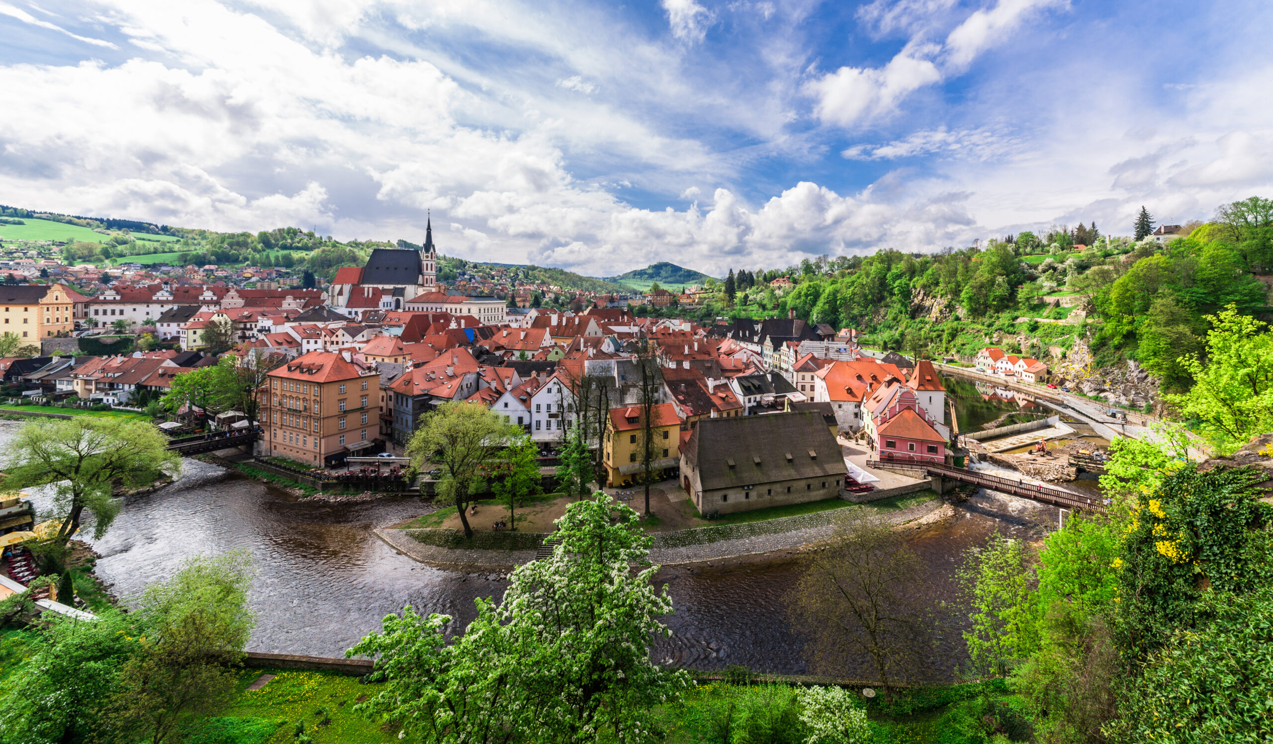 Panoramic view of a European town with red-roofed houses along a winding river, a church spire, and green hills under a blue sky.