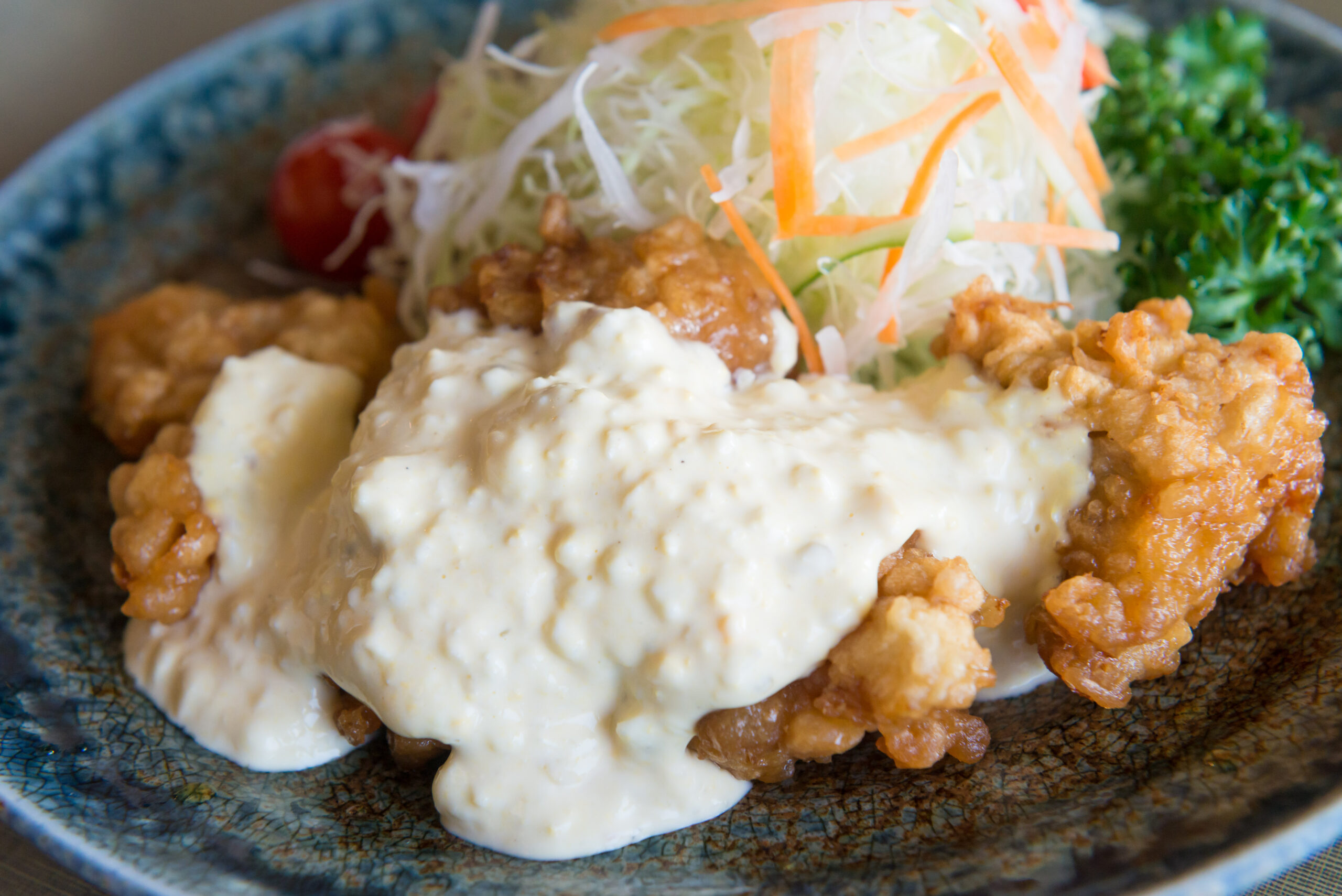 Fried chicken with creamy white sauce on a blue plate, topped with shredded cabbage–carrot slaw and greens in the background.]','Fried chicken with a white sauce on a blue ceramic plate, alongside shredded cabbage and carrot salad.