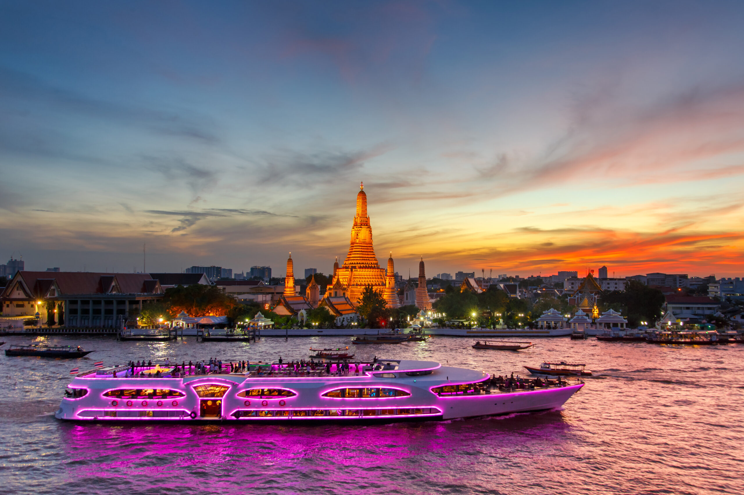Sunset over Bangkok featuring Wat Arun's central spire and a purple-lit river cruise boat in the foreground.