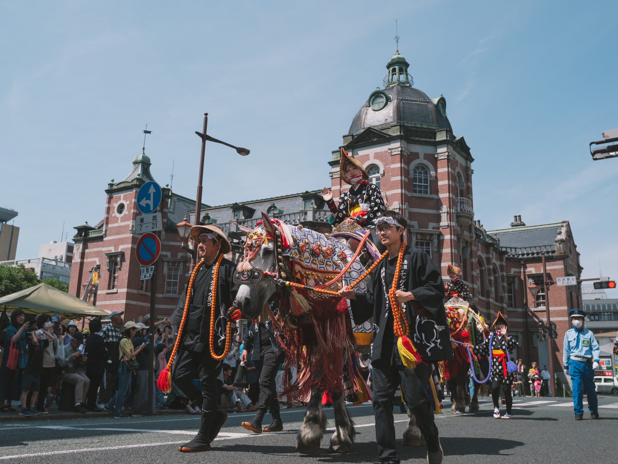 Riders in traditional black outfits on ornate-decorated horses parade down a city street with spectators and a red-brick historic building in the background.