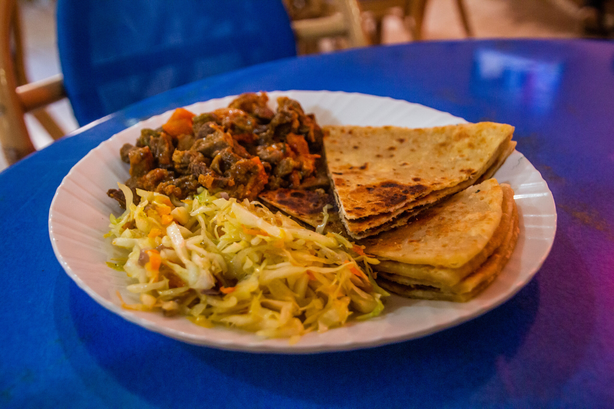 Plate with cooked vegetables, beans, and shredded cabbage beside stacked flatbread on a blue table surface.