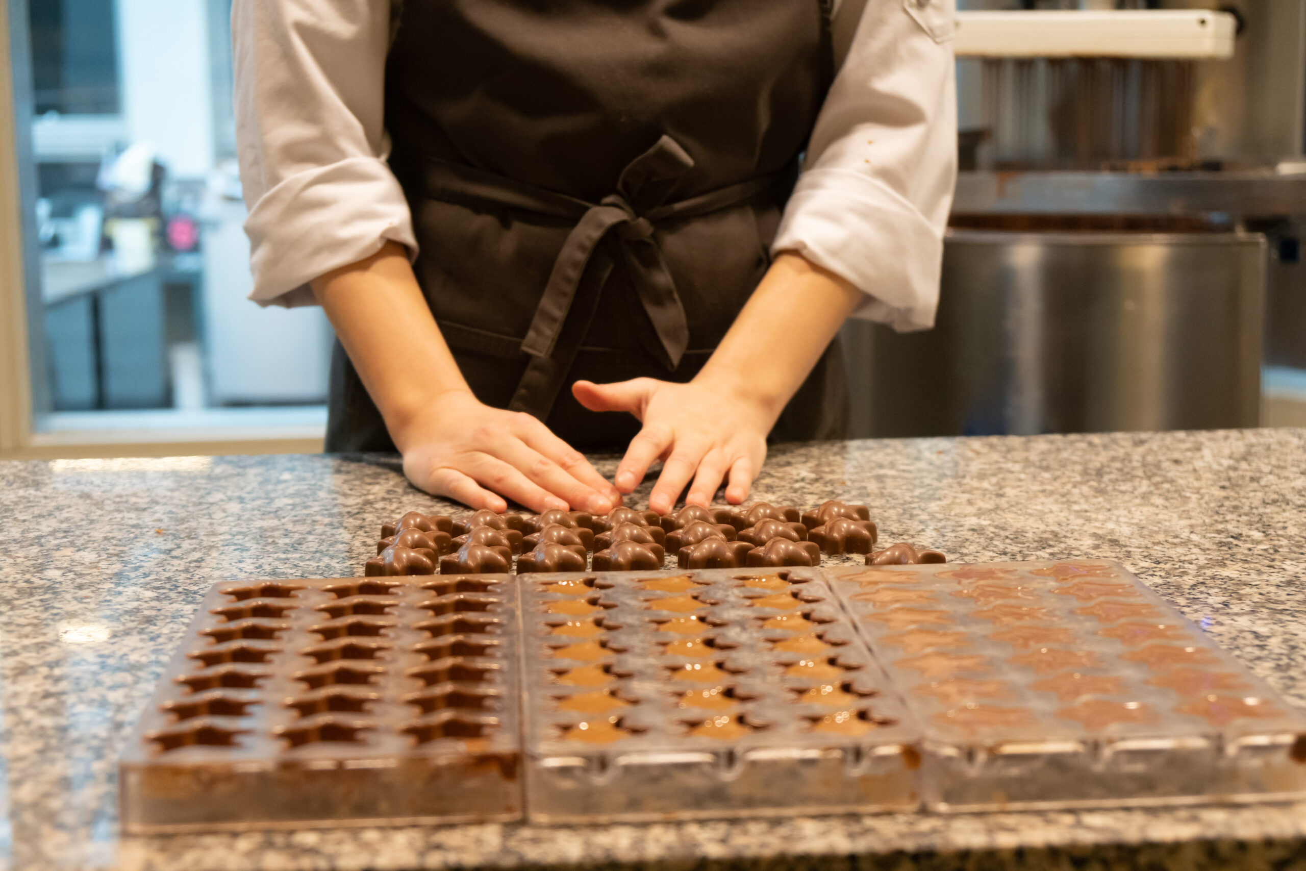 Hands shaping chocolate on a glossy counter, rows of filled chocolate molds nearby in a kitchen.
