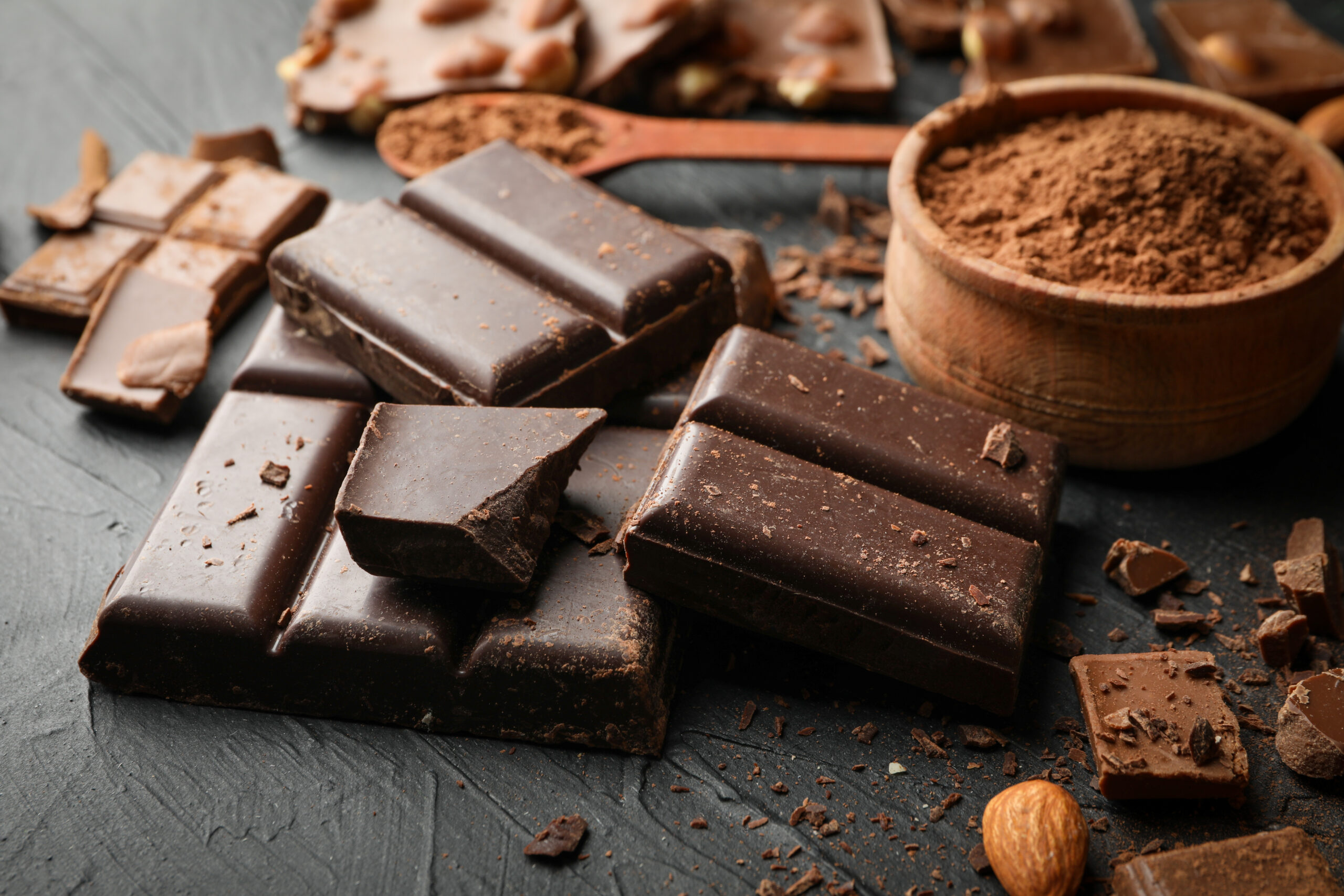 Assorted dark chocolate bars on a slate surface with a bowl of cocoa powder and scattered cocoa dust and nuts in the background.