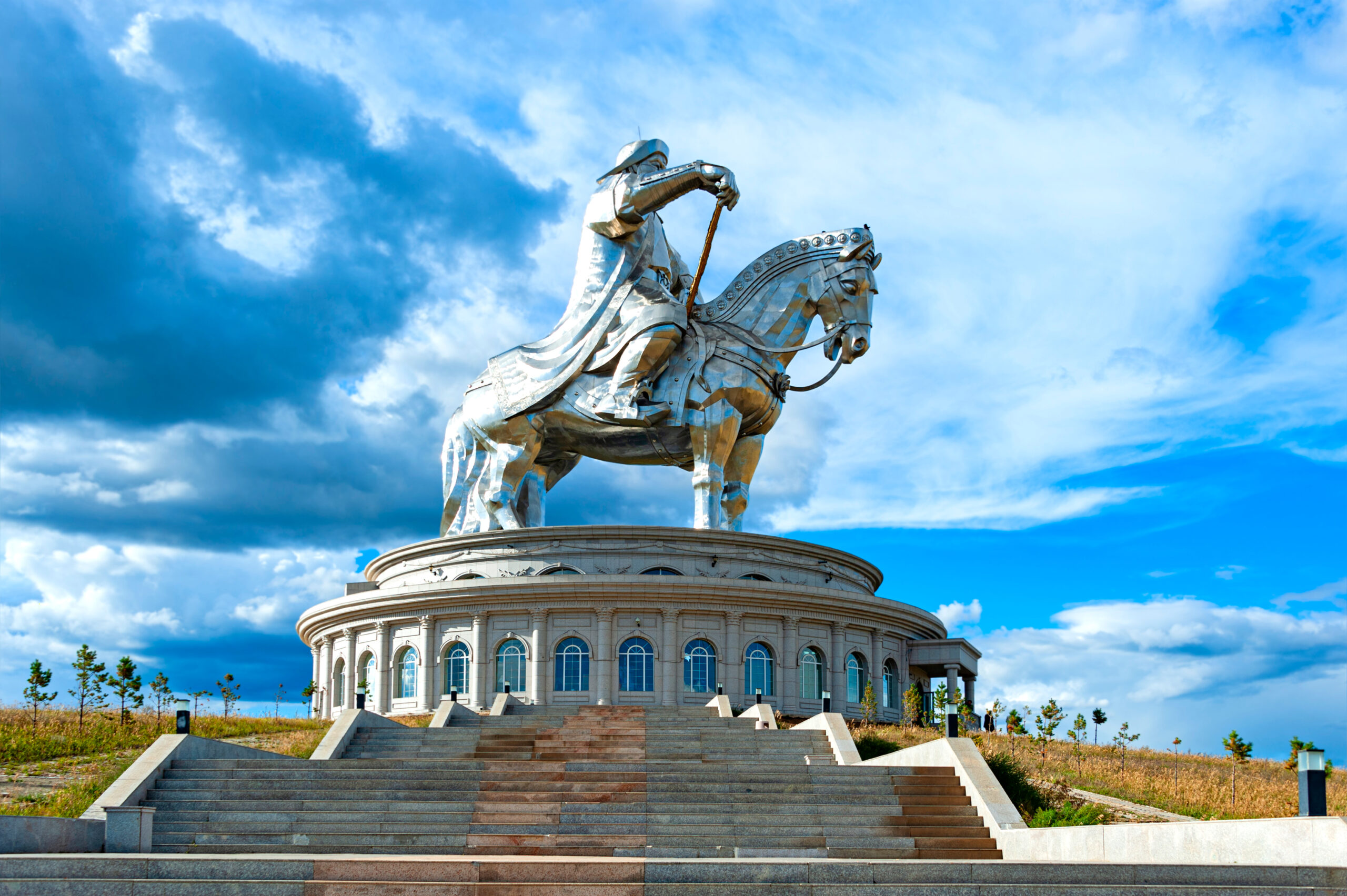 Large silver equestrian statue of a rider on a horse atop a circular pedestal with steps, under a blue cloudy sky.