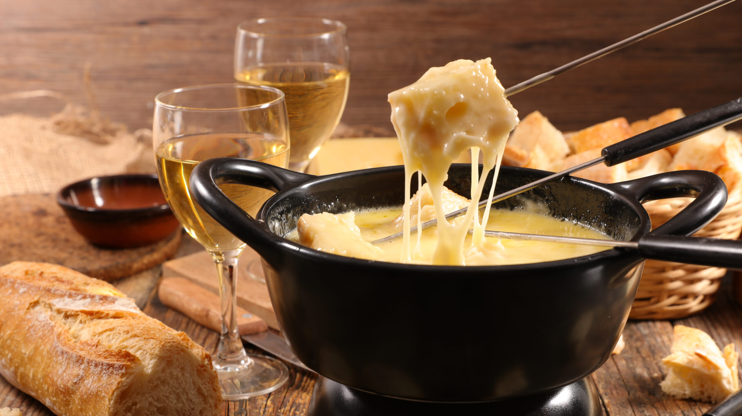 A black fondue pot with melted cheese being dipped by a skewer, surrounded by bread chunks and two glasses of white wine on a wooden table.