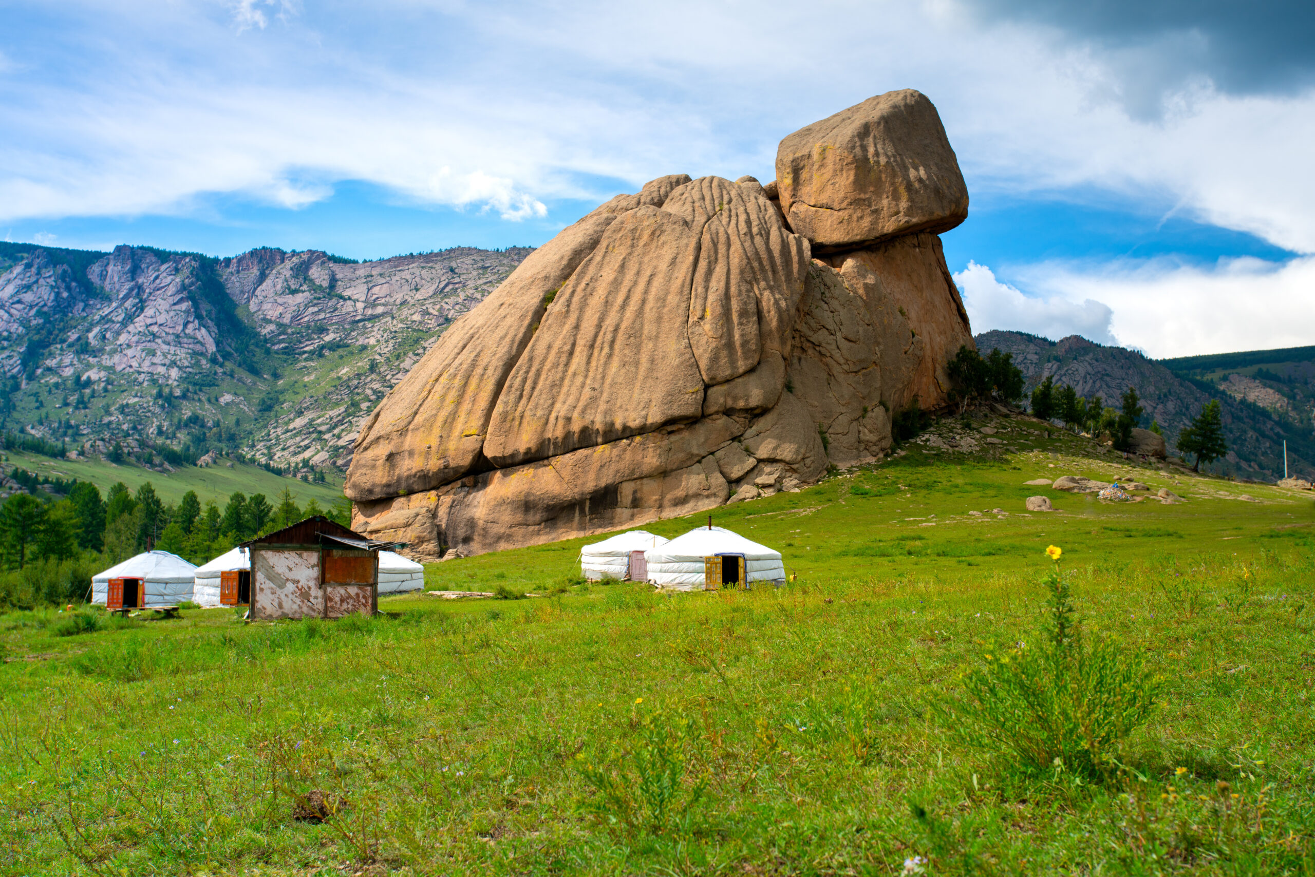 Large sandstone rock formation in a grassy valley with white yurts and small huts at its base, mountains in the distance under a blue sky.