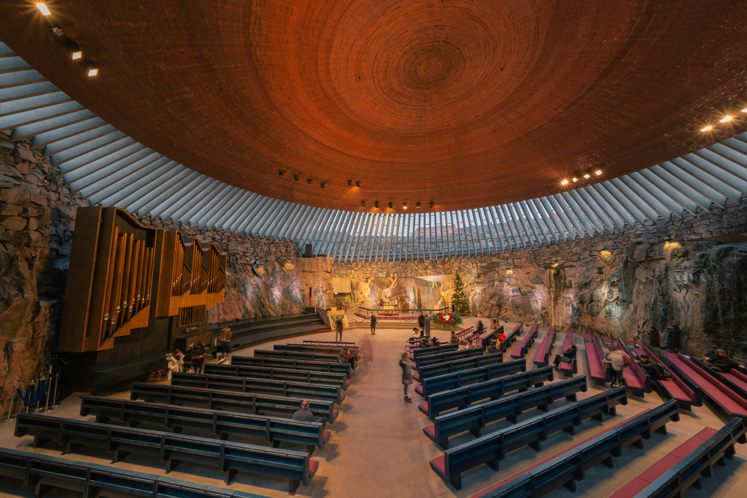 Interior of a circular stone auditorium with a warm wooden domed ceiling, organ on the left, and tiered seating filled with a few visitors.