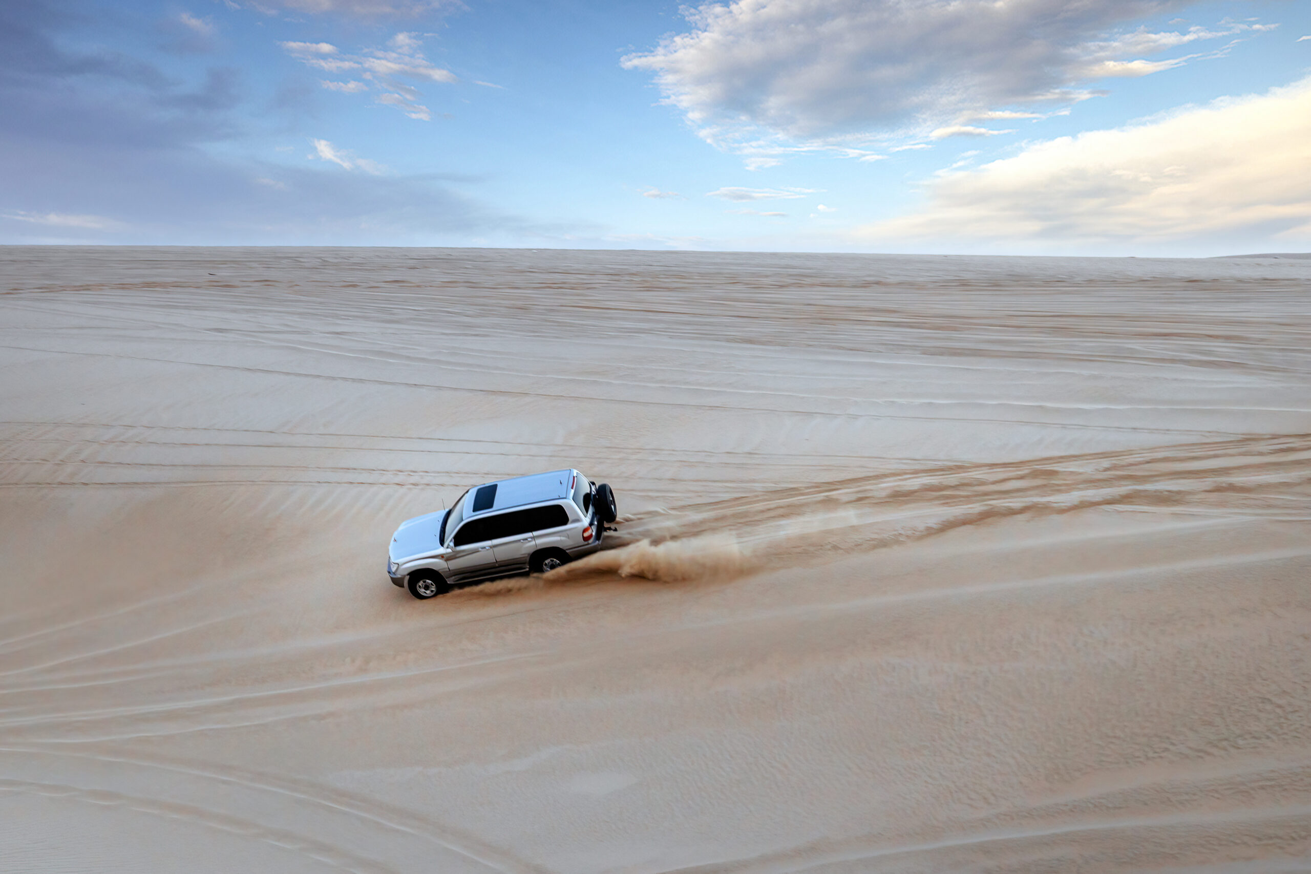 Silver SUV skidding down a sandy desert slope with a wide blue sky above.