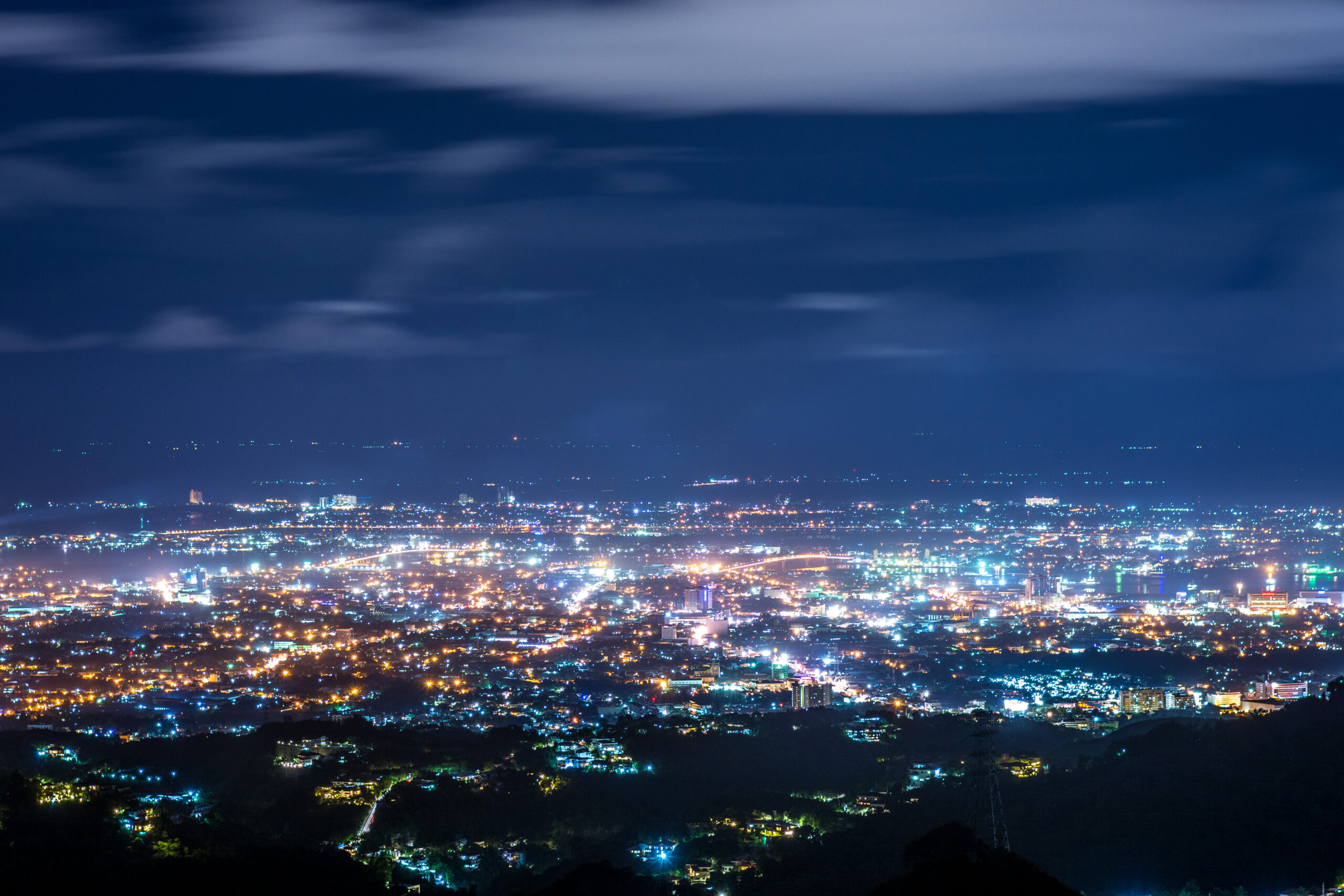 Distant city lights glow below a dark blue night sky with wispy clouds and a silhouetted hillside in the foreground.