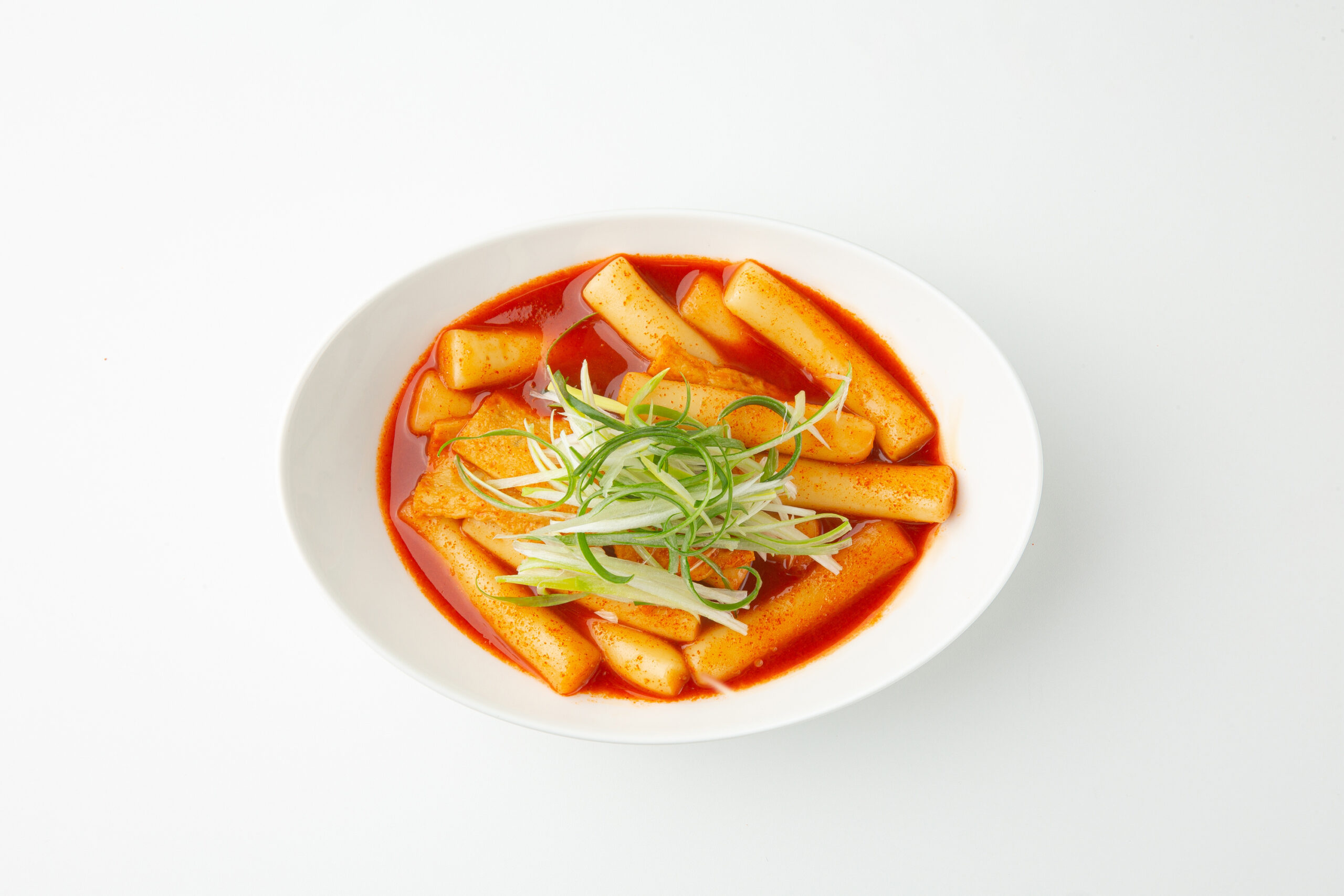 Bowl of spicy red broth with cylindrical white rice cakes and green onion garnish on top.