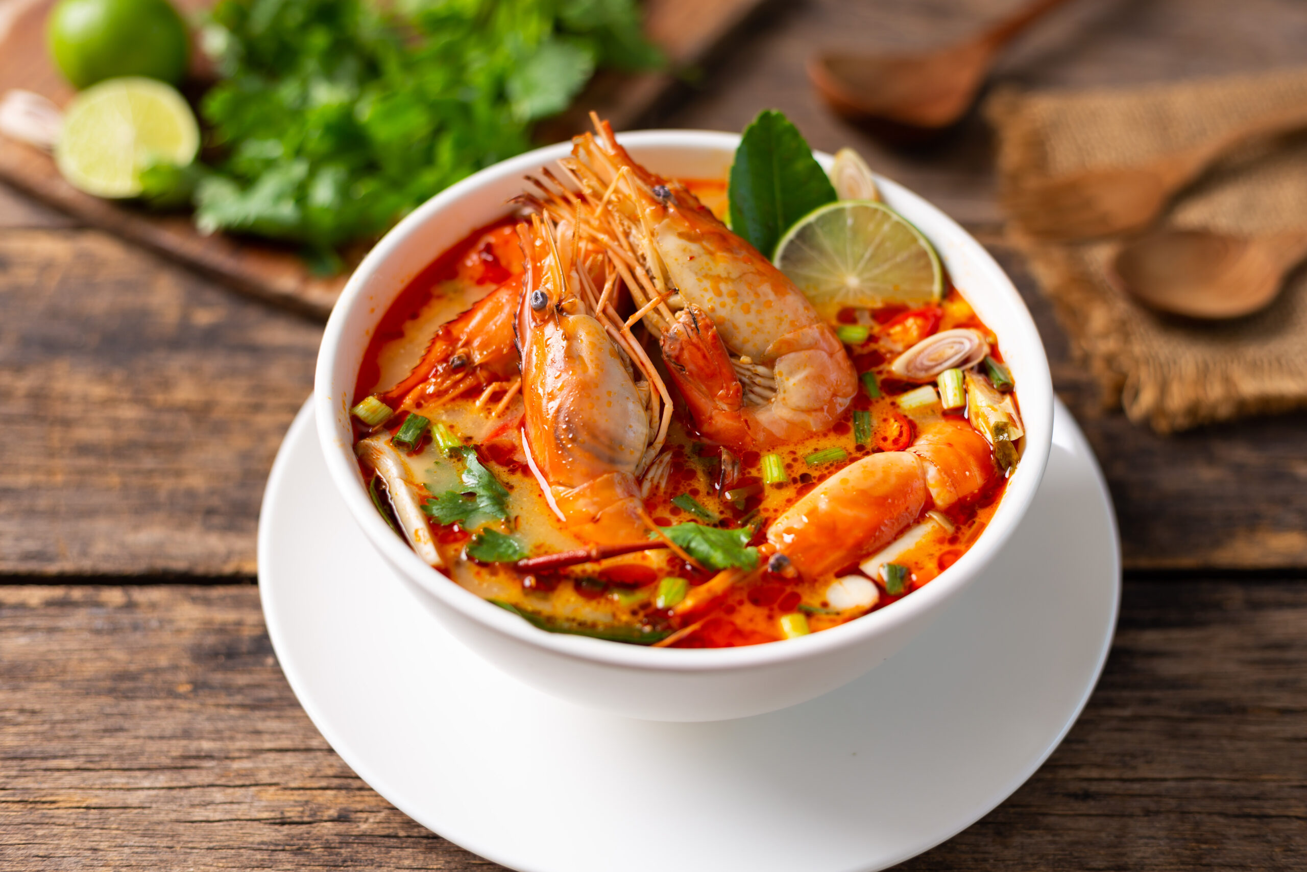 Bowl of spicy seafood soup with large shrimp, lime and herbs on a wooden table.