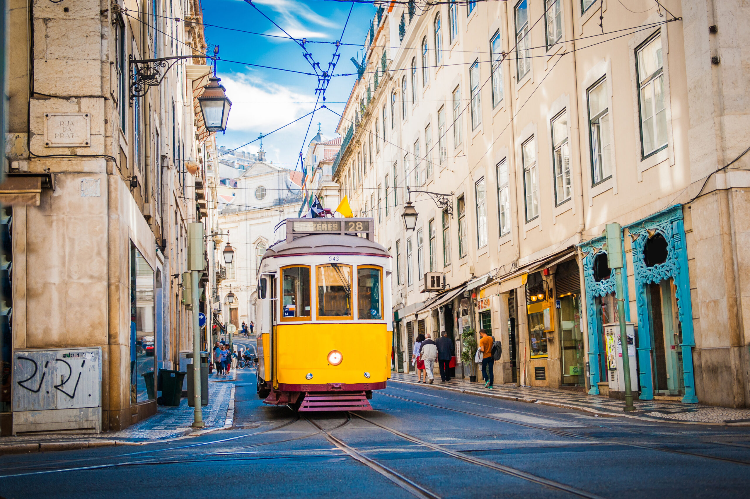 Yellow tram travels down a narrow street lined with tall pastel buildings and crisscrossing overhead wires, blue sky above.