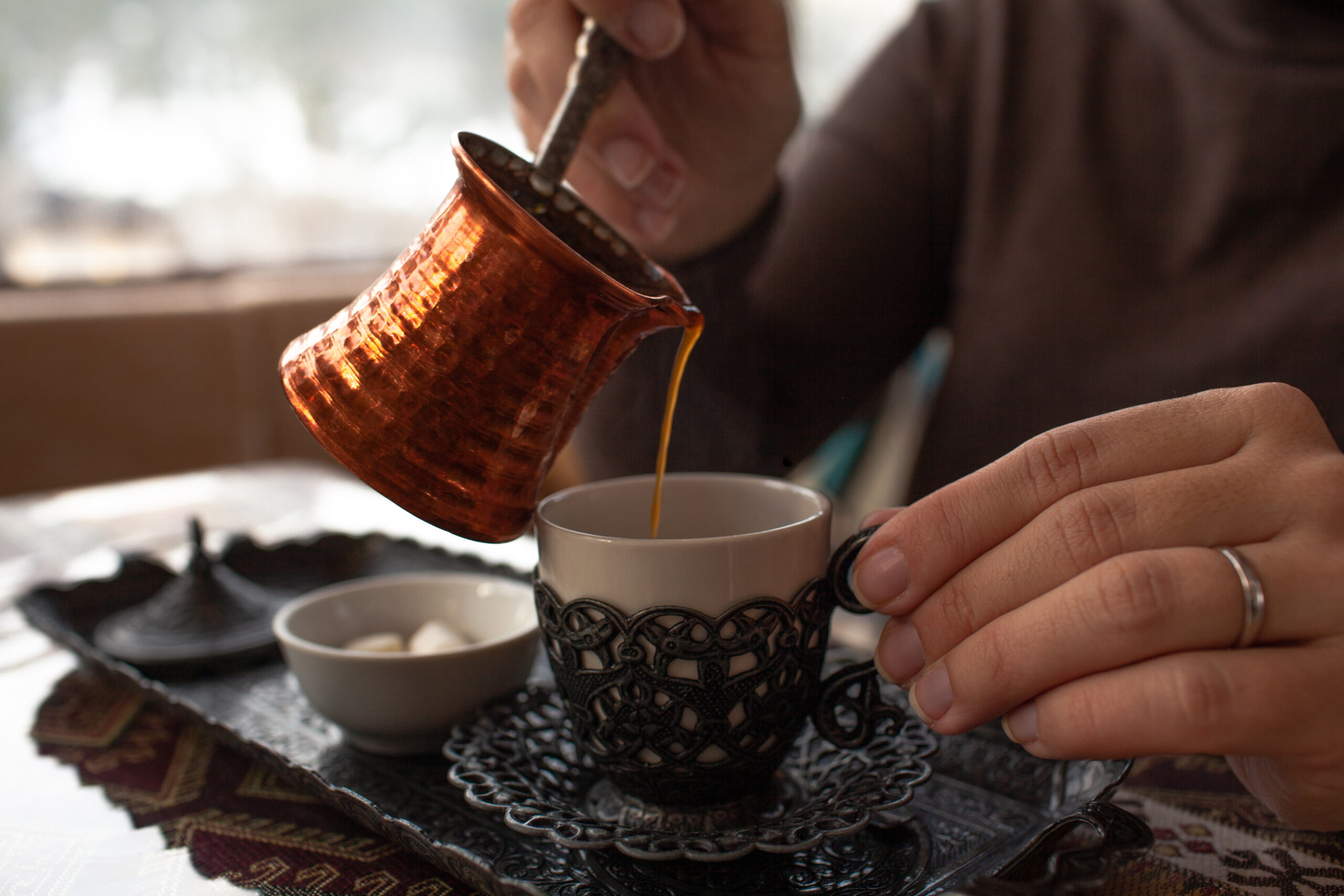 Person pours hot coffee from a copper cezve into a white cup on a lace table, with hands steady nearby.