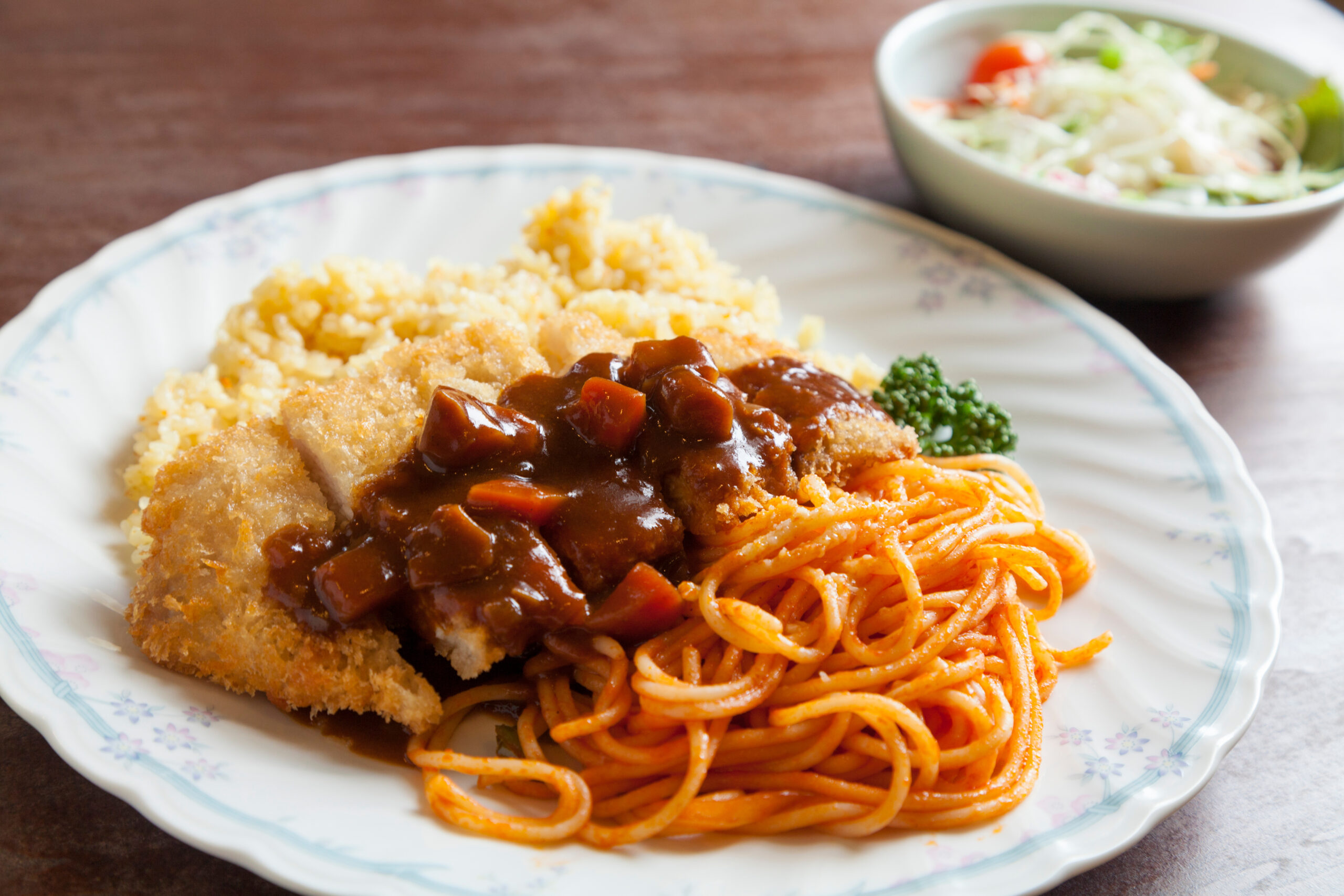 Plate with a breaded fried cutlet covered in brown gravy, alongside yellow fried rice, broccoli, and spaghetti with tomato sauce; blurred salad bowl in the background.