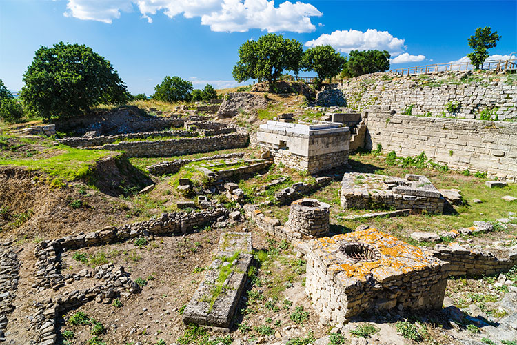 Ancient ruins with stone walls and foundations on grassy, sunlit site under a blue sky with trees nearby